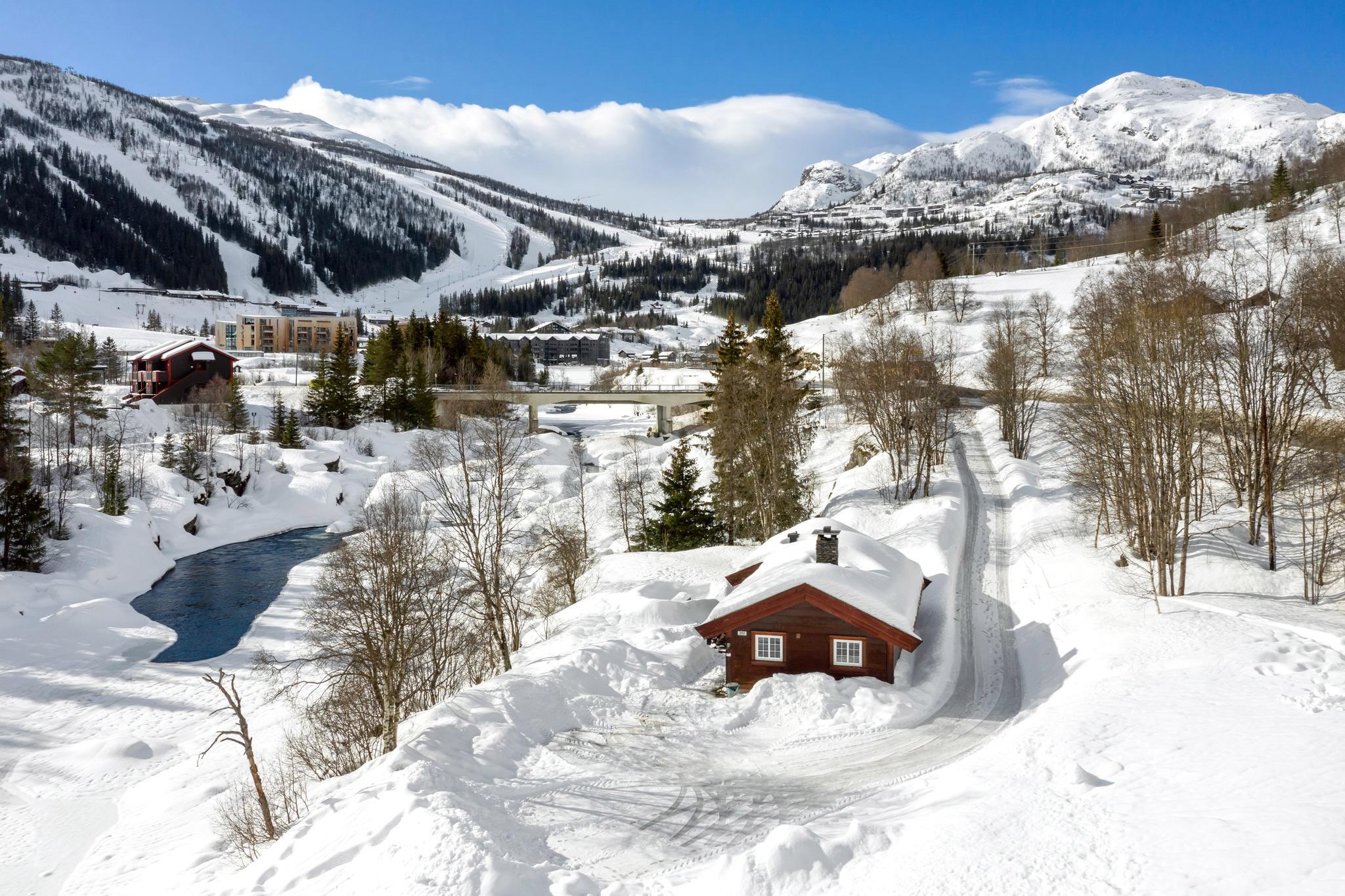 Et snelandskab byder på en hyggelig træhytte omgivet af snedækkede træer, perfekt til en skiferie. En lille flod løber ved siden af, med bjerge, der minder om dem i Saalbach i baggrunden under en klar blå himmel. En vej fører til hytten, omkranset af høje, snefyldte træer.