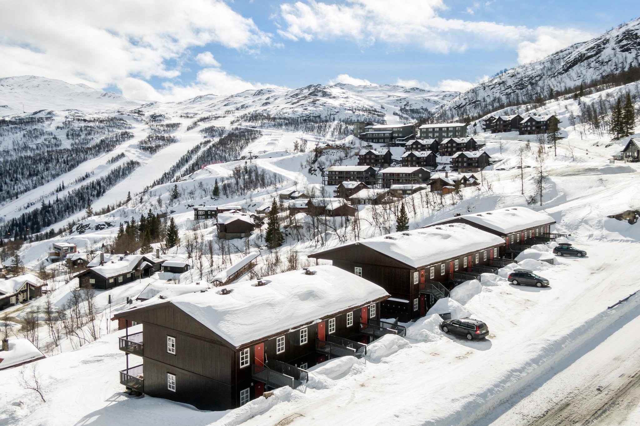 Snedækket bjerglandskab med mørke træhytter og parkerede biler skaber en charmerende scene i Westendorf. En snoet vej og skiløjper er synlige i baggrunden. Den lyseblå himmel med spredte skyer tilføjer kontrast til dette maleriske vinterlandskab, ideelt for skirejser.