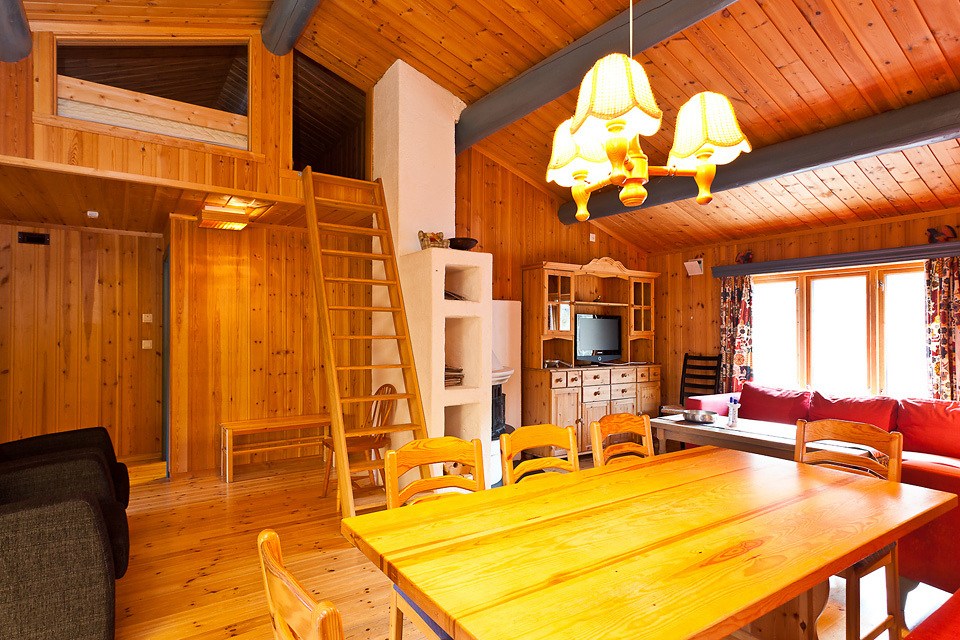 En hyggelig kabine af træ i Westendorf har et spisebord og stole i forgrunden. En hems med stige er på den ene side, mens et opholdsområde med rød sofa og tv ved et stort vindue vækker charmen ved sommerferie Østrig. Varm belysning fremhæver de rustikke funktioner.