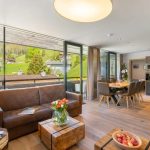 Lyst, moderne opholds- og spiseområde med store vinduer, der viser en naturskøn udsigt over bjergsiden, der minder om Saalbach. Værelset har en brun sofa, et sofabord i træ med blomster, et spisebord til seks personer og et åbent tekøkken. Varm belysning og indretning fremhæver den indbydende atmosfære.