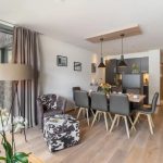 Moderne opholds- og spisestue i åbent plan med trægulv og stort vindue, perfekt til at minde om en skiferie i Saalbach. Et spisebord med otte stole er sat til et måltid. Der er en mønstret lænestol, gulvlampe og vægmonterede fotos. Køkken i baggrunden.
