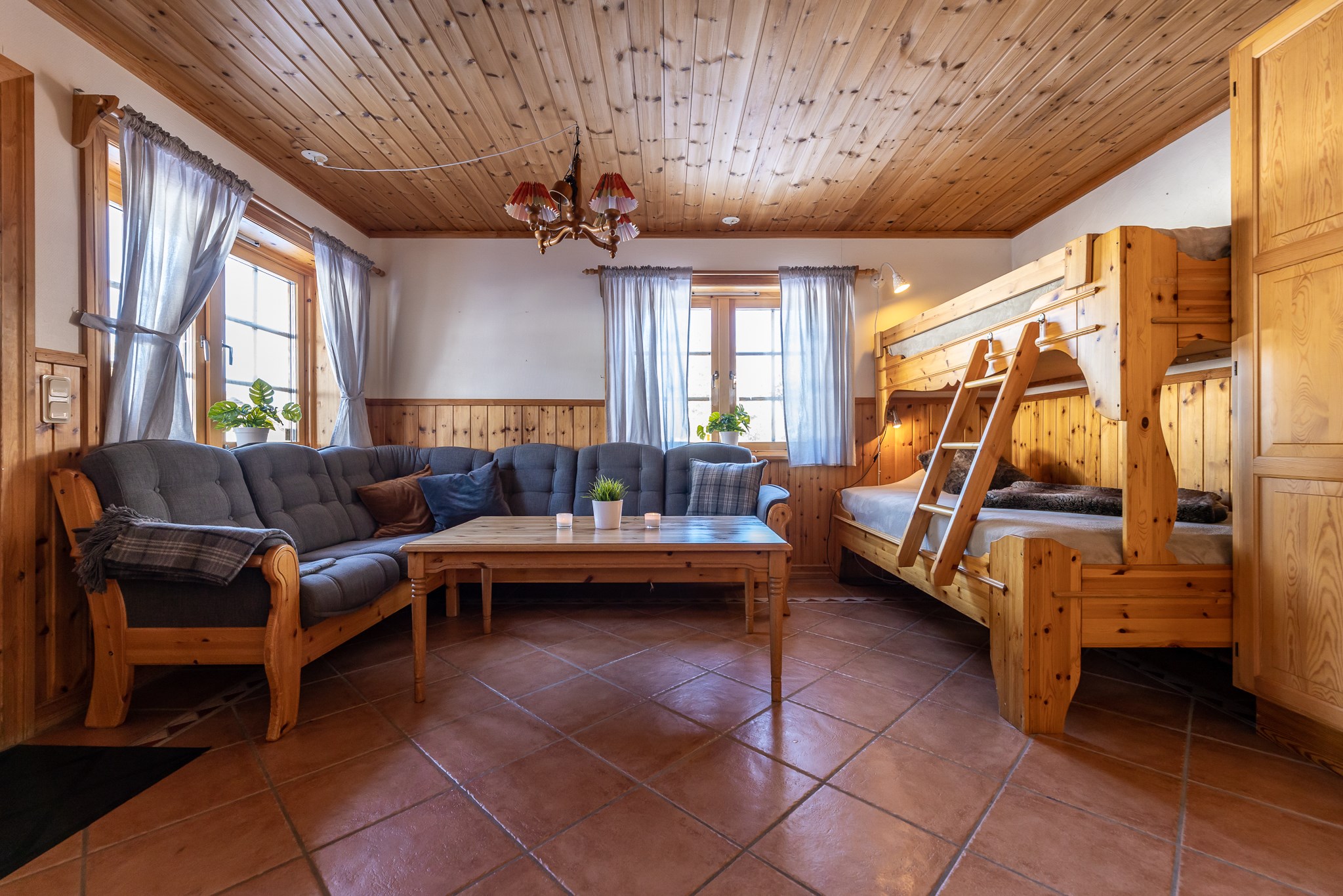En hyggelig stue med trævægge og -loft, der minder om et Westendorf-retræte, har en stor L-formet sofa, et sofabord i træ, en rustik lysekrone og køjesenge. To vinduer med gennemsigtige gardiner slipper naturligt lys ind, mens klinkegulve tilføjer et varmt touch.