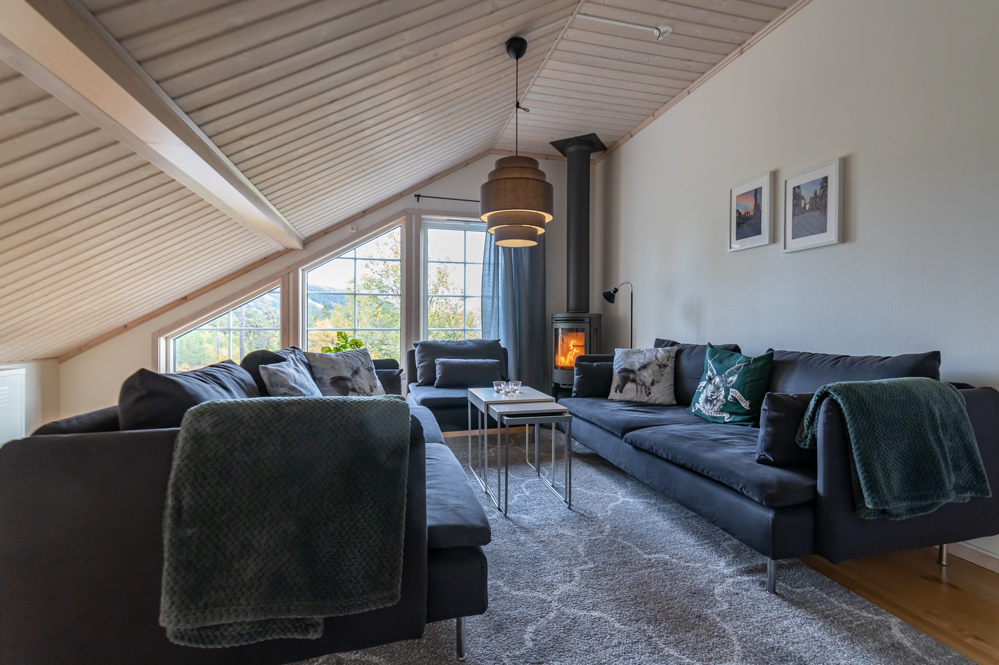 En hyggelig stue i Westendorf med et skrånende træloft har to mørke sofaer, et sofabord og en pejs. Store vinduespartier til venstre lukker naturligt lys ind. Indretningen inkluderer to indrammede billeder og en vævet loftslampe. Et plystæppe dækker gulvet, perfekt efter skirejseeventyr.