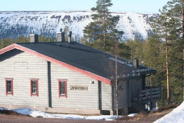 En træhytte med sort tag og rødbeklædte vinduer ligger omgivet af træer i Westendorf. Et skilt ved siden af vinduerne lyder Vinterro. Et snedækket bjerg er synligt i baggrunden under en klar blå himmel, der tilbyder den perfekte skiferie flugt.