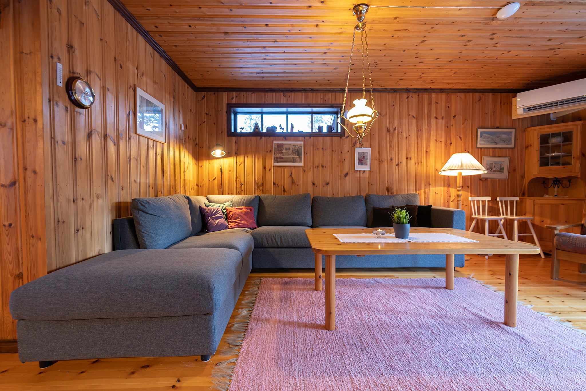 En hyggelig stue med trævægge og loft, der minder om en skiferie i Westendorf, har en stor grå sektionssofa, et sofabord i træ på et lyserødt tæppe og flere varme lamper. En lille plante pryder bordet, og indrammede billeder pryder væggene.