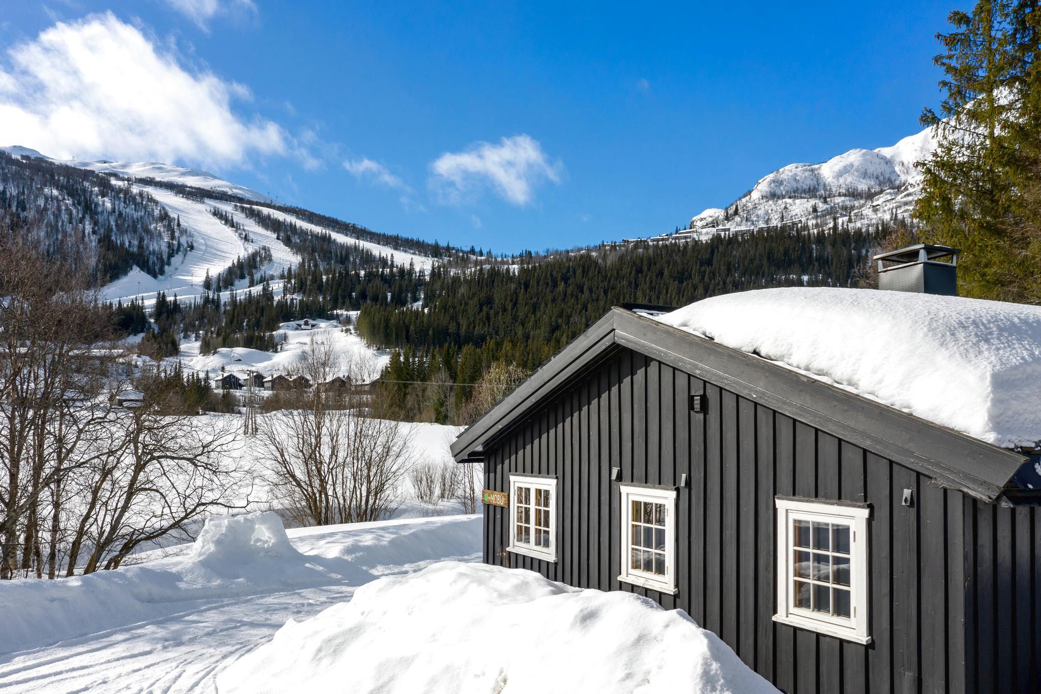 En hyggelig sort hytte med snedækket tag, beliggende i Saalbachs vinterlandskab. Sne dækker jorden og træerne, mens skiløjper og skovklædte bjerge strækker sig ind i den klare blå himmel, hvilket gør det til et ideelt sted for en skiferie.