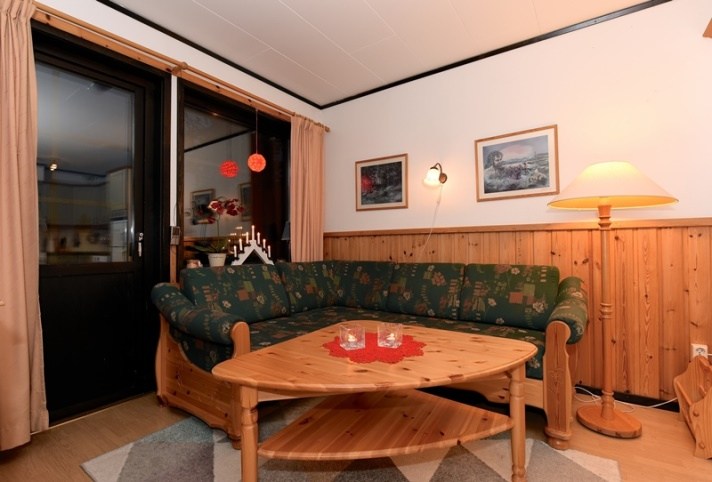 En hyggelig stue med en mørkegrøn mønstret sofa og et sofabord i træ fremkalder charmen ved Westendorf. Væggene, dels træpaneler dels beige, er prydet med landskabsmalerier, der minder om en sommerferie Østrig. En høj gulvlampe og et gardinvindue fuldender den festlige scene.