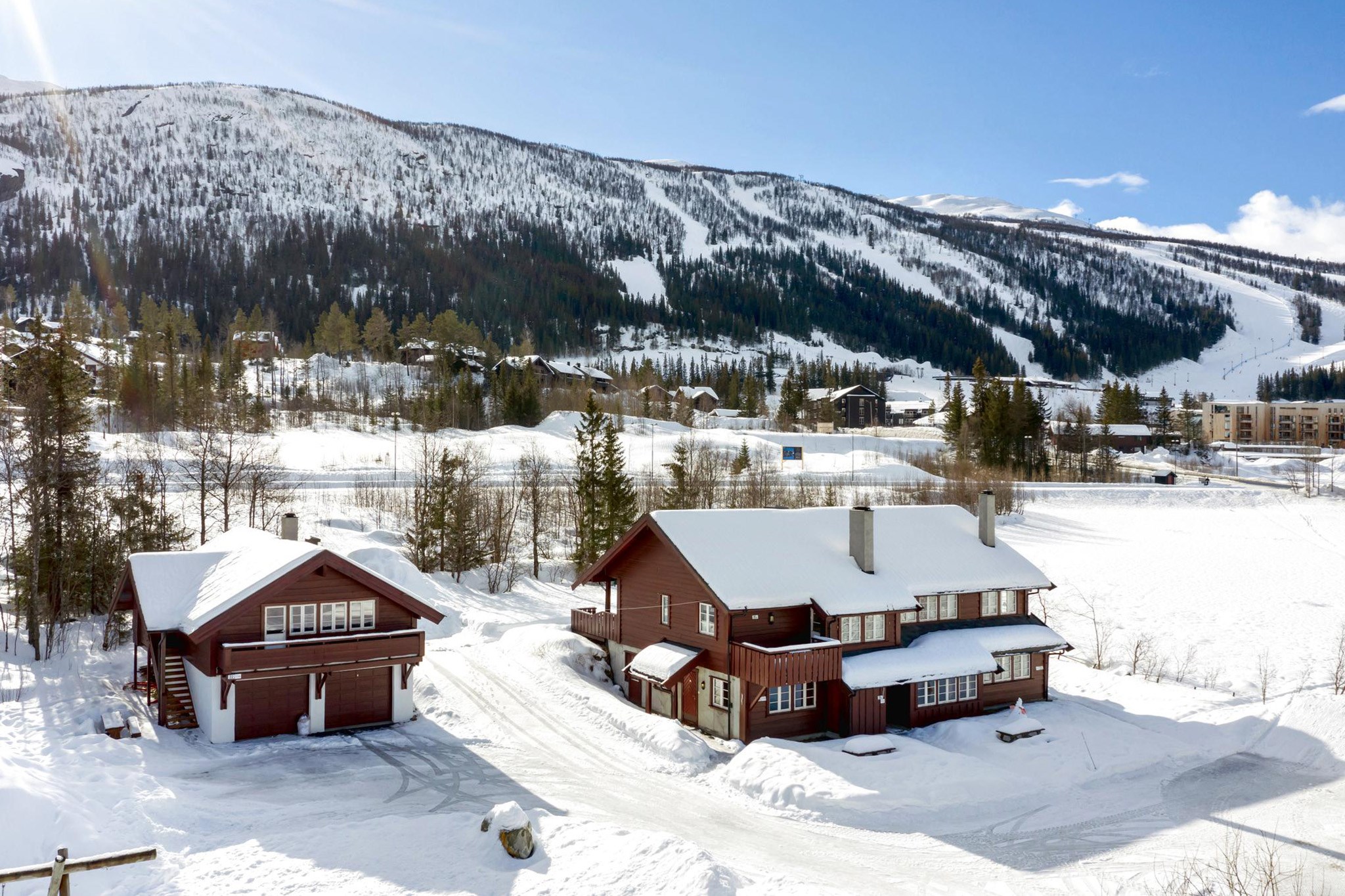To træhytter med snedækkede tage er beliggende i et snelandskab, omgivet af træer og bakker. En klar blå himmel og bjergbaggrunden i Wagrain fuldender denne naturskønne vinterudsigt, perfekt til en skiferie-flugt.