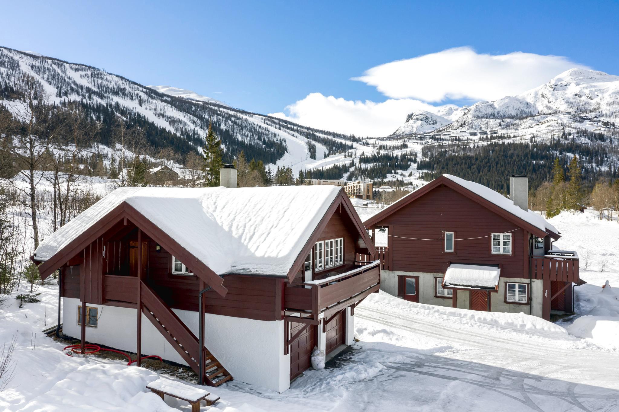 To træhytter med snedækkede tage står i et snelandskab, der minder om en fredfyldt skiferie. I baggrunden møder de sneklædte bjerge i Saalbach en knaldblå himmel. En skiløjpe snor sig gennem bjerglandskabet og byder på spændende eventyr ligesom i Westendorf.