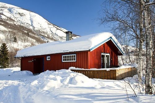 En rød hytte med snedækket tag ligger midt i dyb sne og nøgne træer og fremkalder charmen ved en skiferie i Saalbach. I baggrunden tårner et solbelyst bjerg sig op under en klar blå himmel og byder på en malerisk scene for vinterentusiaster.