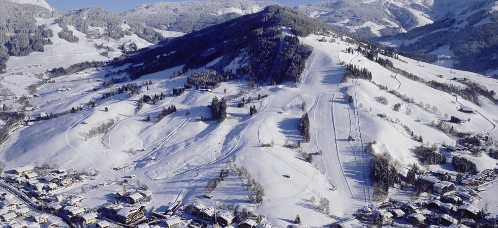 Luftfoto af et snedækket bjerglandskab med Saalbachs skisportssted. Talrige skiløjper er synlige. Nedenfor ligger en landsby med snestøvede hustage i dalen. Himlen er klar og lyseblå, hvilket forstærker denne vinterlige skiferiescene.