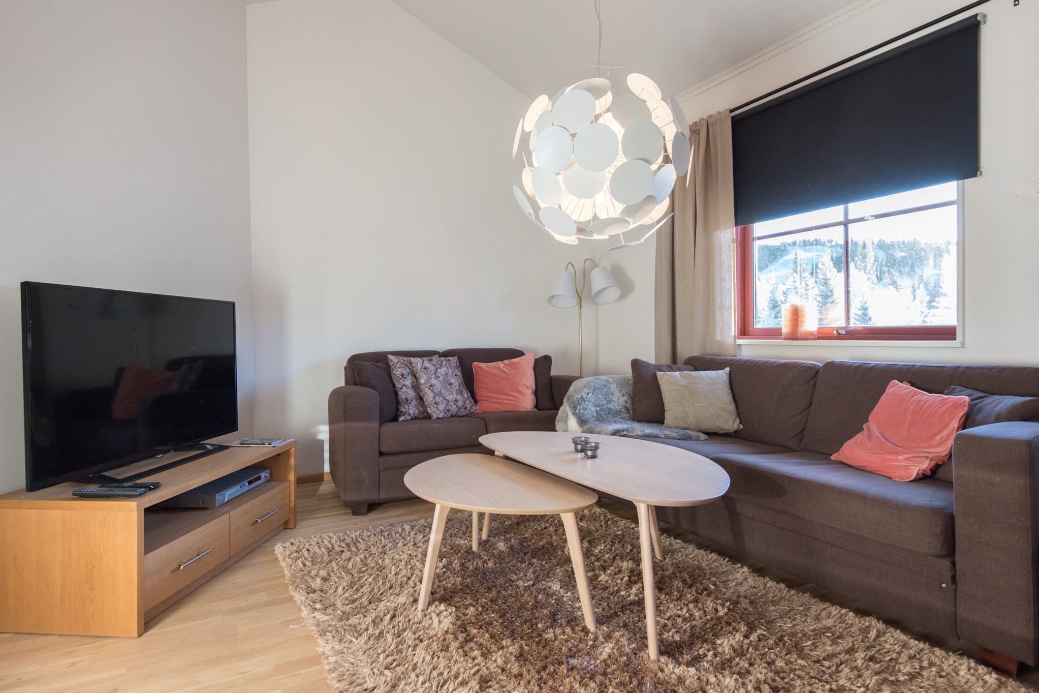 En hyggelig stue med en brun sofa og lænestol, dekoreret med lyserøde og grå puder, vækker minder om et Saalbach-retræte. Et rundt sofabord i træ sidder på et plys tæppe. Et stort tv står på en træstativ. Fra loftet hænger en moderne pendel, og et vindue viser en sneklædt udsigt, der minder om skirejseeventyr.