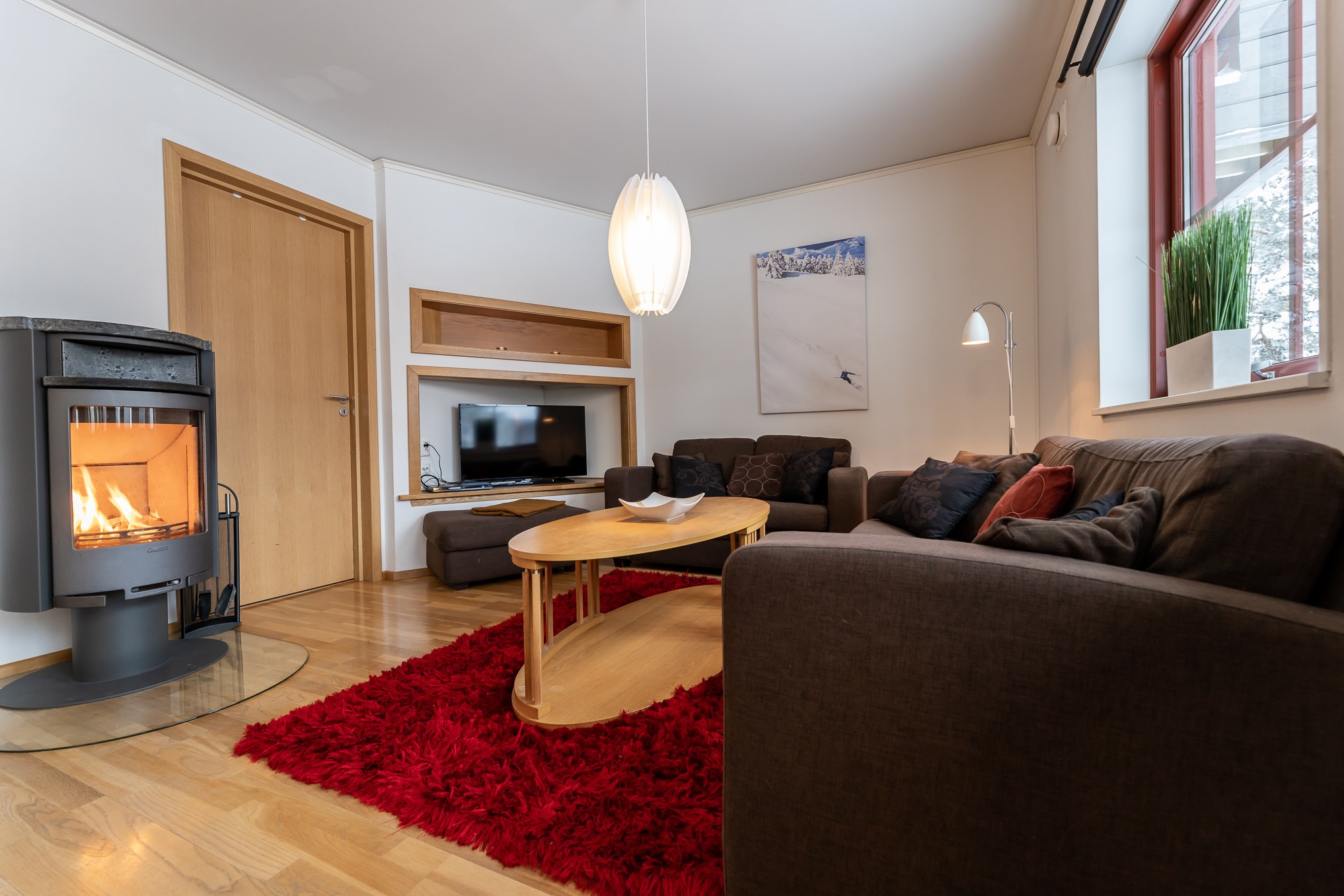 En hyggelig stue med et moderne design udstråler varme som en hytte i Wagrain. Der er en tændt pejs, fladskærms-tv og brune sofaer omkring et sofabord i træ på et rødt shag tæppe. Et plante- og snelandskabsbillede tilføjer indretning, mens et stort vindue lysner rummet op.