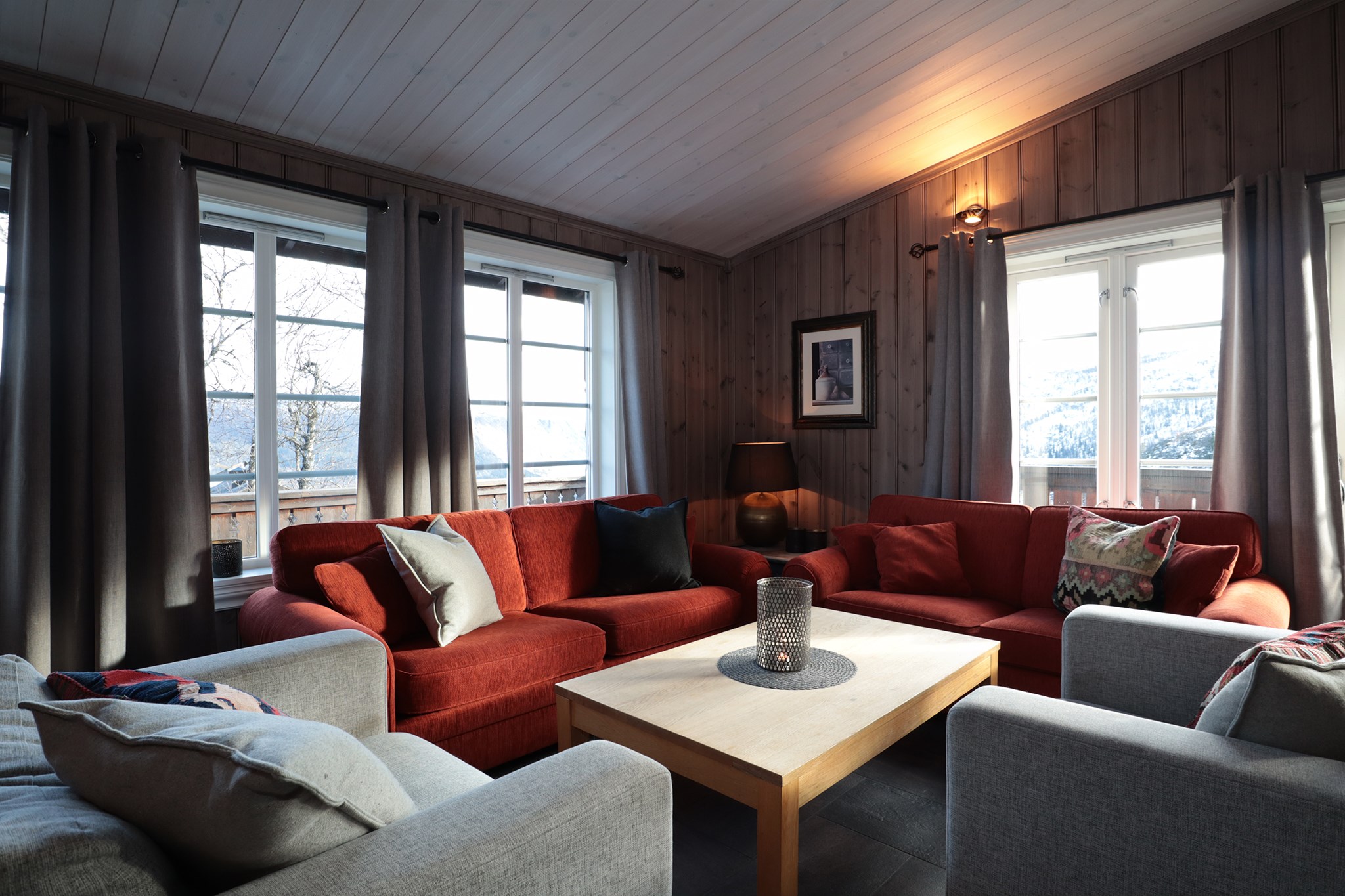 En hyggelig stue i Westendorf har røde sofaer og grå lænestole omkring et sofabord af træ. Store vinduespartier med grå gardiner slipper naturligt lys ind. En lille lampe på et sidebord oplyser rummet, og et billede hænger på den træbeklædte væg.