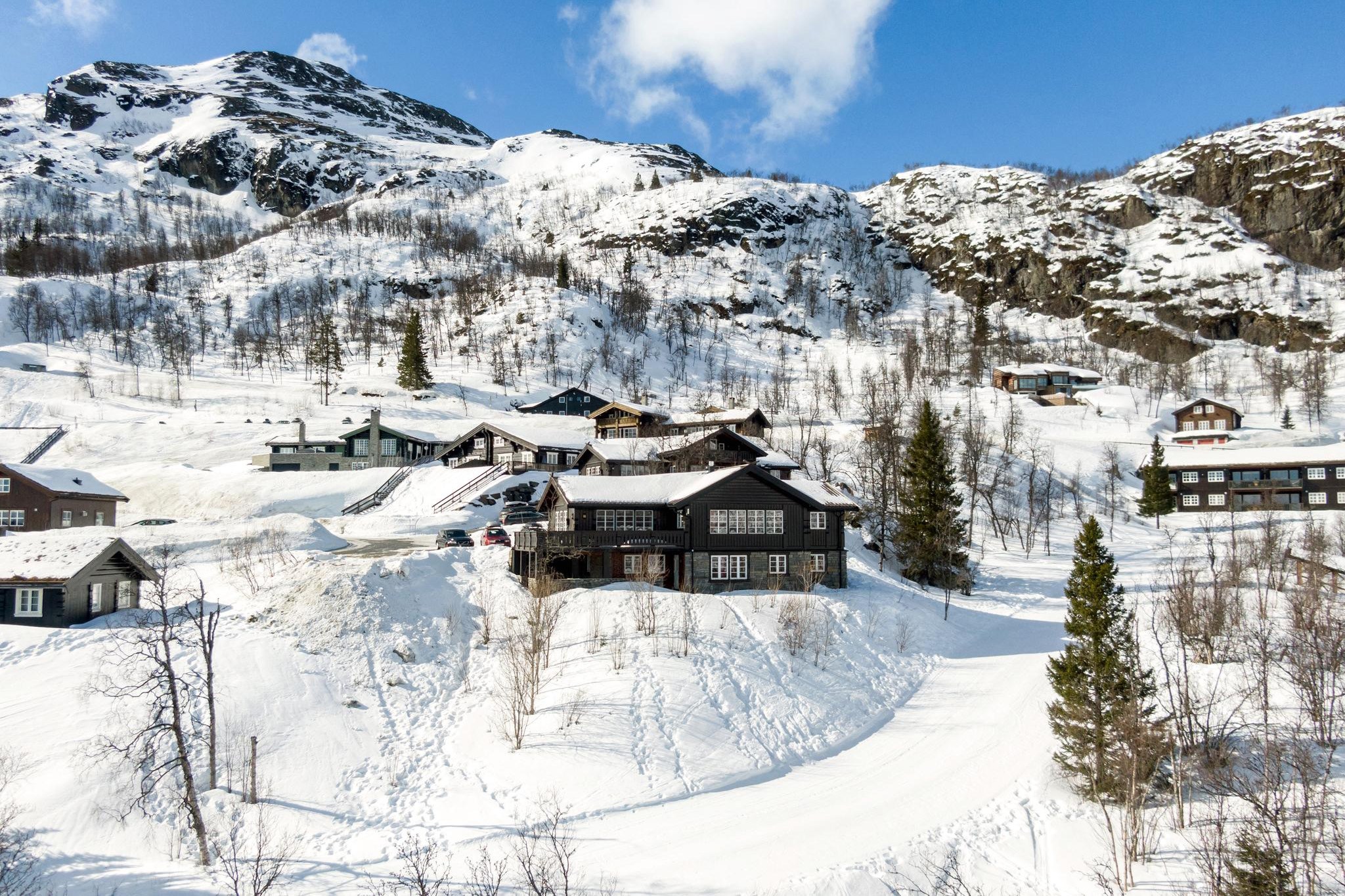 Et sneklædt bjerglandskab i Westendorf byder på flere træhytter spredt på en bjergskråning. Området er dækket af sne, med skiløjper synlige i forgrunden. Fyrretræer er spredt overalt, og klippeklipper rejser sig i baggrunden under en klar blå himmel.