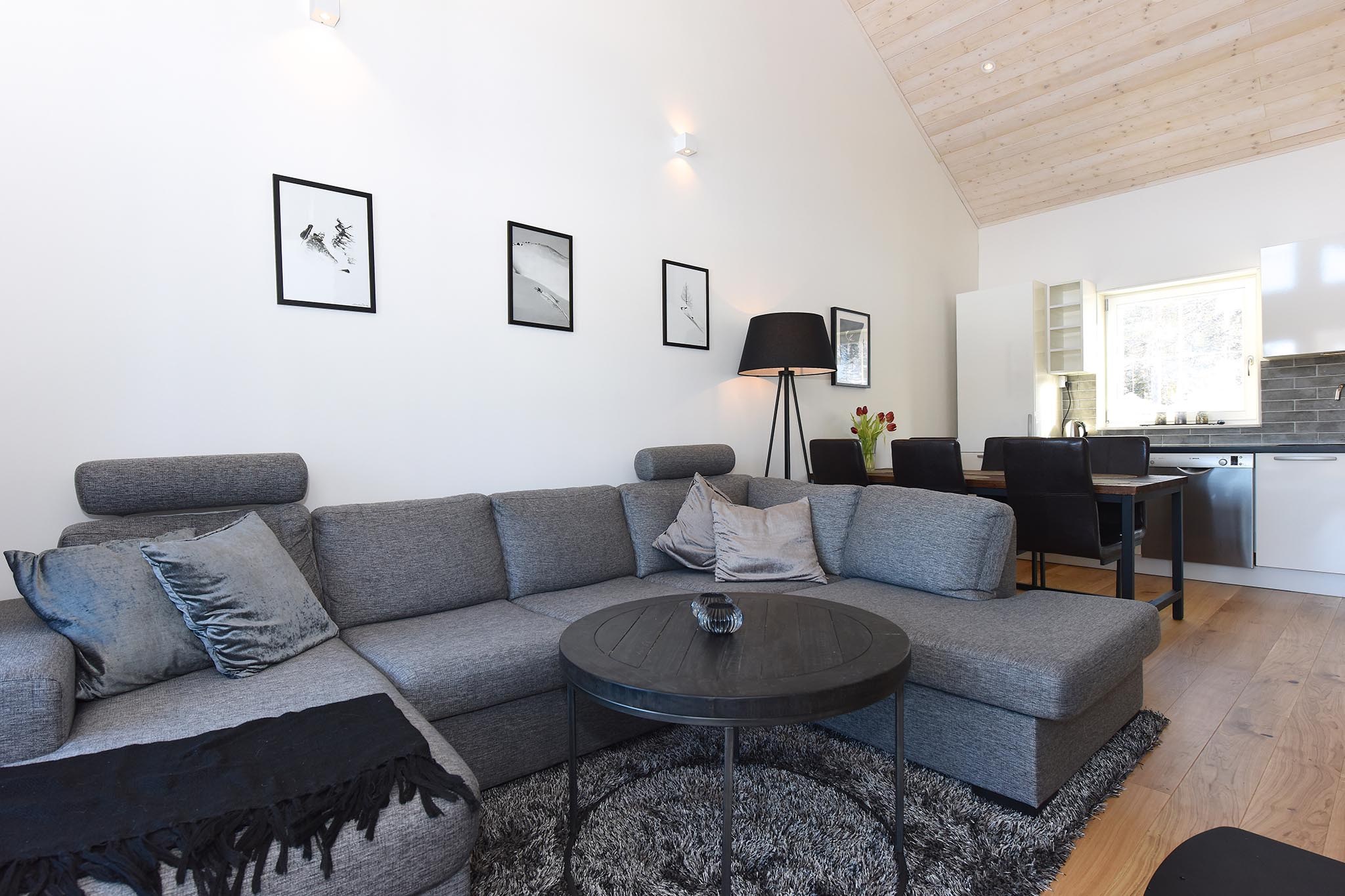 En moderne stue har en grå sektionssofa på et rundt tæppe, suppleret med en sort gulvlampe og indrammet kunst. Et rundt sofabord sidder foran sofaen. Spisepladsen og køkkenet er synlige med trægulve overalt, der minder om den hyggelige charme, der findes i Westendorf.
