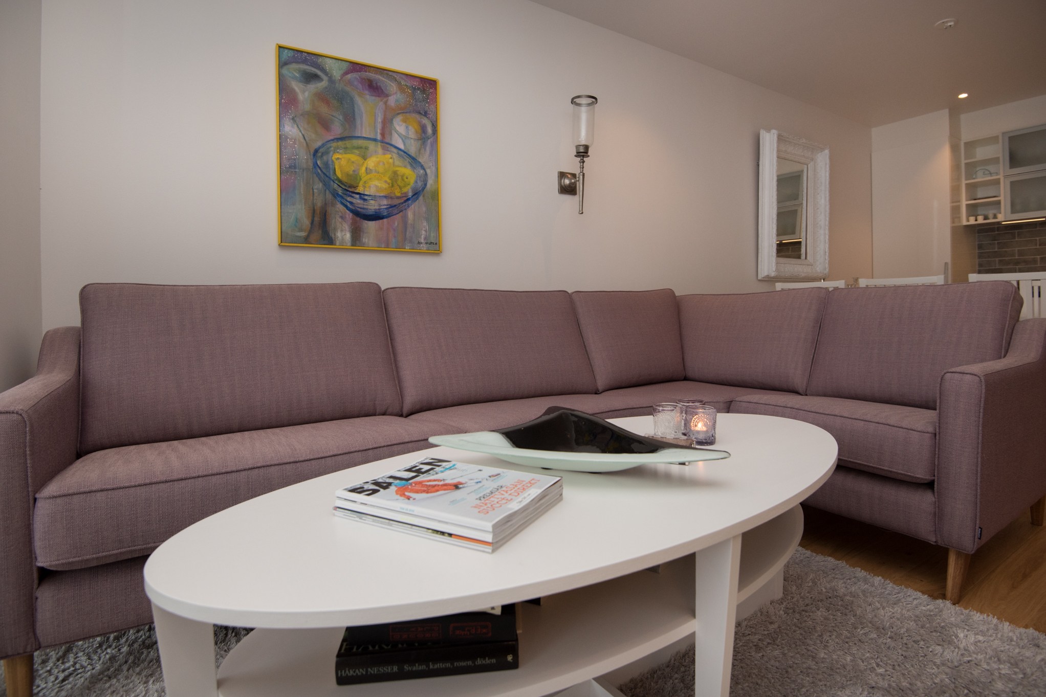 En hyggelig stue med en lilla L-formet sofa, et hvidt ovalt sofabord og dekorative genstande fremkalder Westendorfs fredfyldte charme. Et levende abstrakt maleri og spejl pryder væggen. Værelset har trægulve og et blødt gråt tæppe, mens stearinlys og magasiner hviler på bordet.