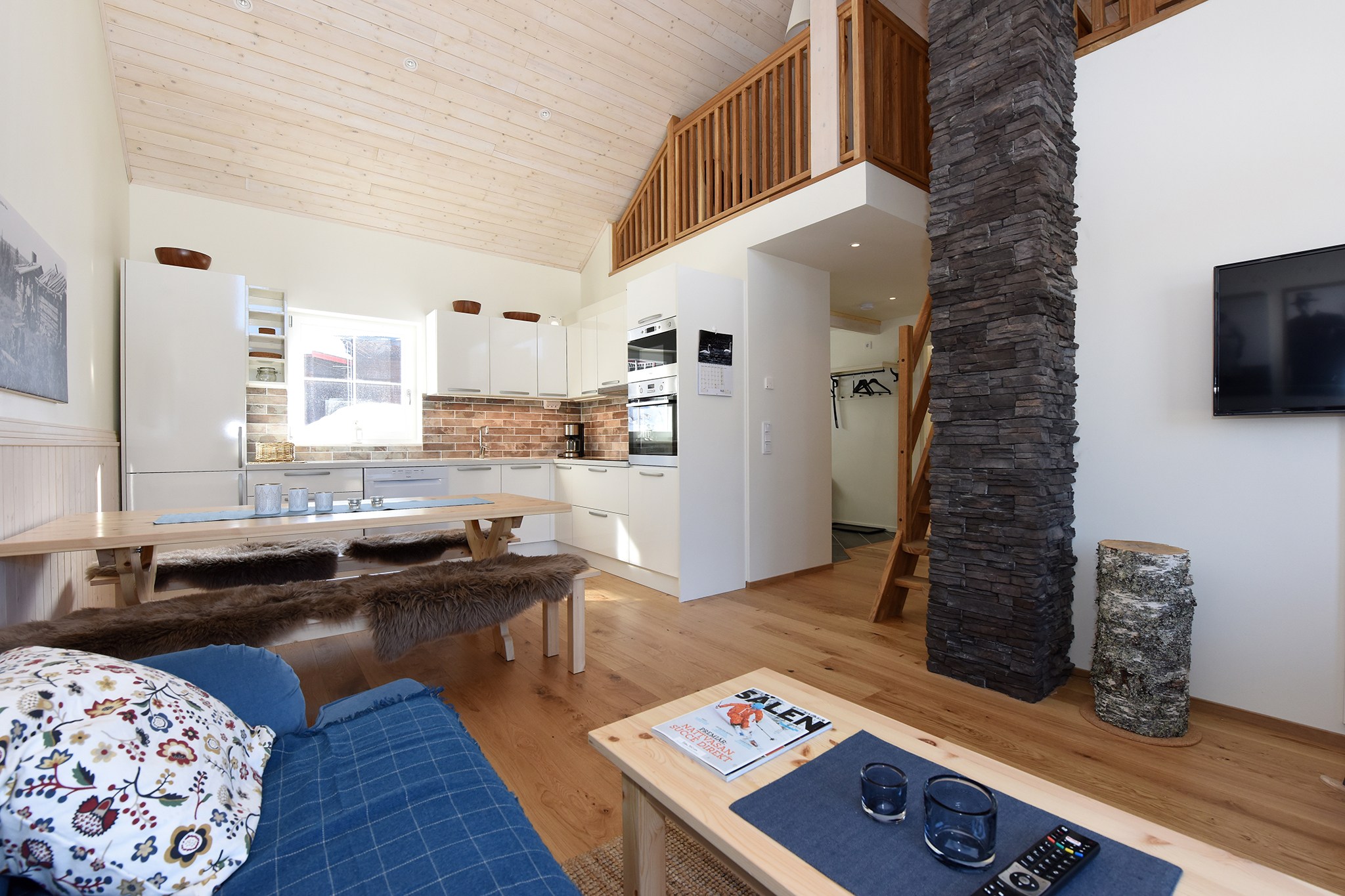 Et hyggeligt opholdsrum i stil med et Saalbach-retræte, med træloft og trægulv. Det kan prale af et moderne køkken med hvide skabe, en åben spiseplads, en behagelig blå sofa og en pejs med stendetaljer. Naturligt lys fylder rummet, der minder om en østrigsk sommerferie.