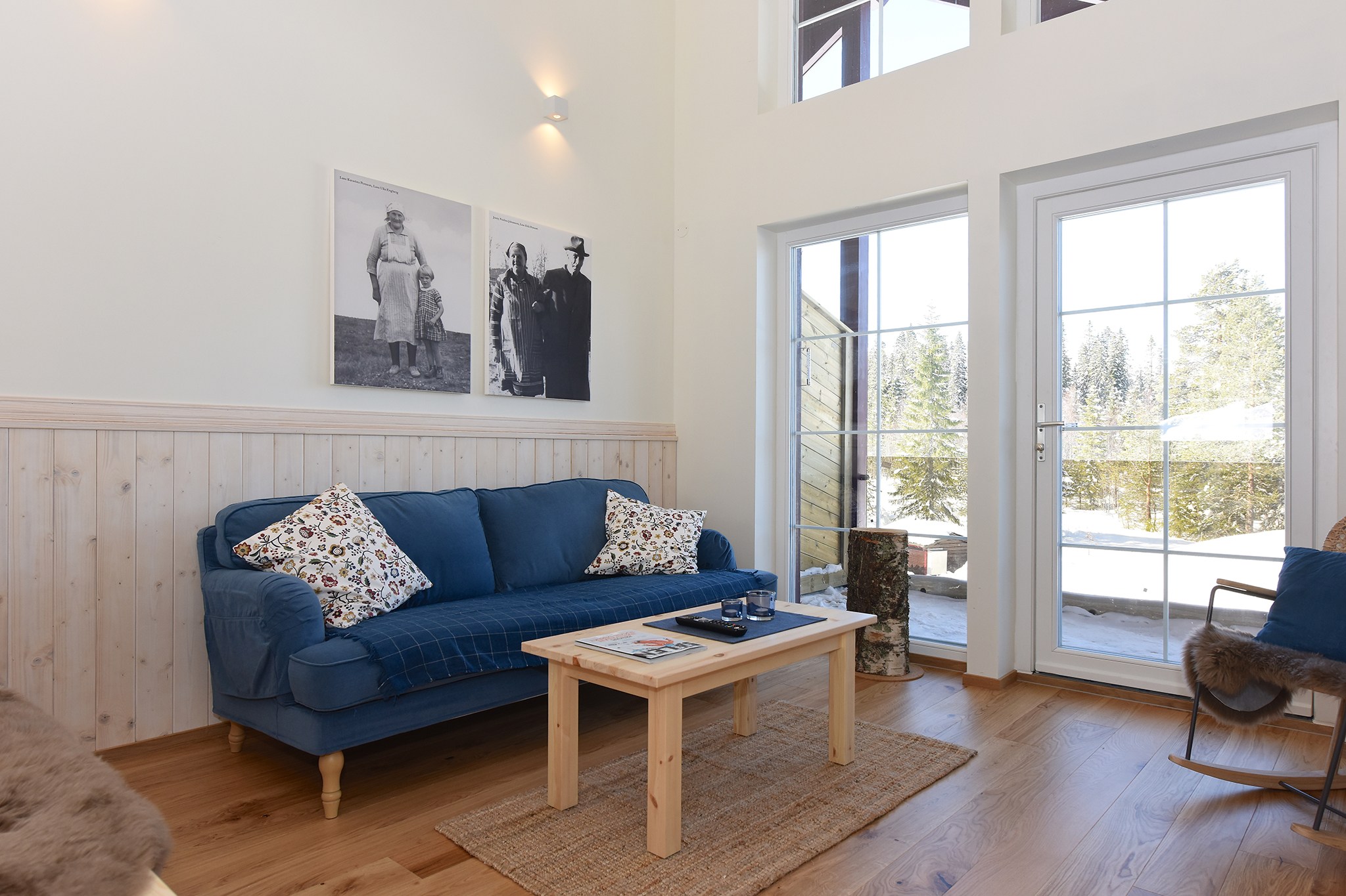 En hyggelig stue med en blå sofa prydet med mønstrede hynder, et sofabord i træ og et stort vindue, der fører til en balkon med udsigt, der minder om Westendorfs charmerende landskaber. Væggene er dekoreret med sort/hvide fotografier. Naturligt lys fylder rummet.