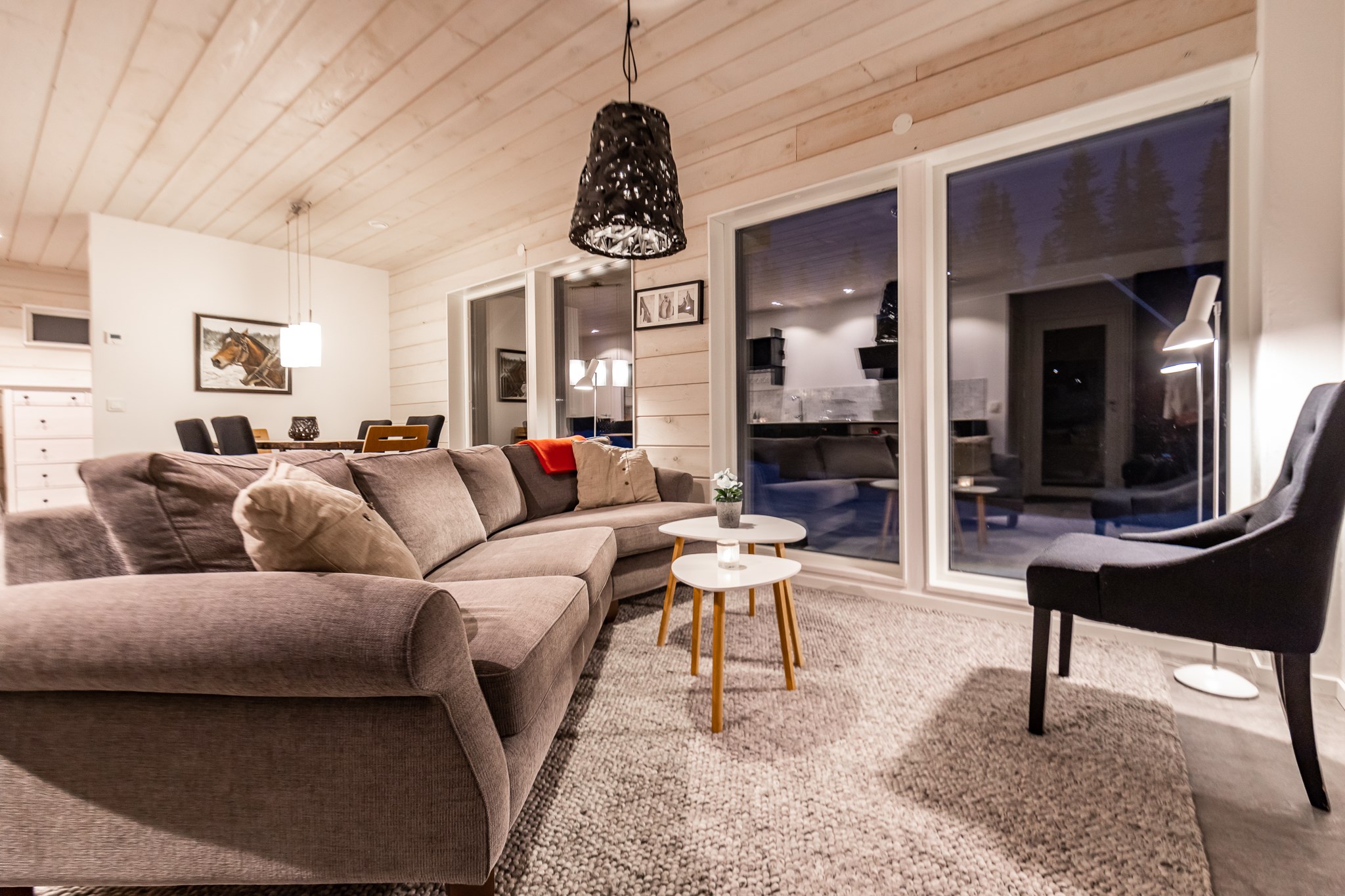 En hyggelig stue med en stor grå sektionssofa, en sort lænestol og et sofabord i træ. Varm belysning, en vævet pendel og gulv-til-loft-vinduer har udsigt over Saalbachs natlige skønhed, hvilket skaber en indbydende atmosfære, perfekt til planlægning af din næste skiferie.