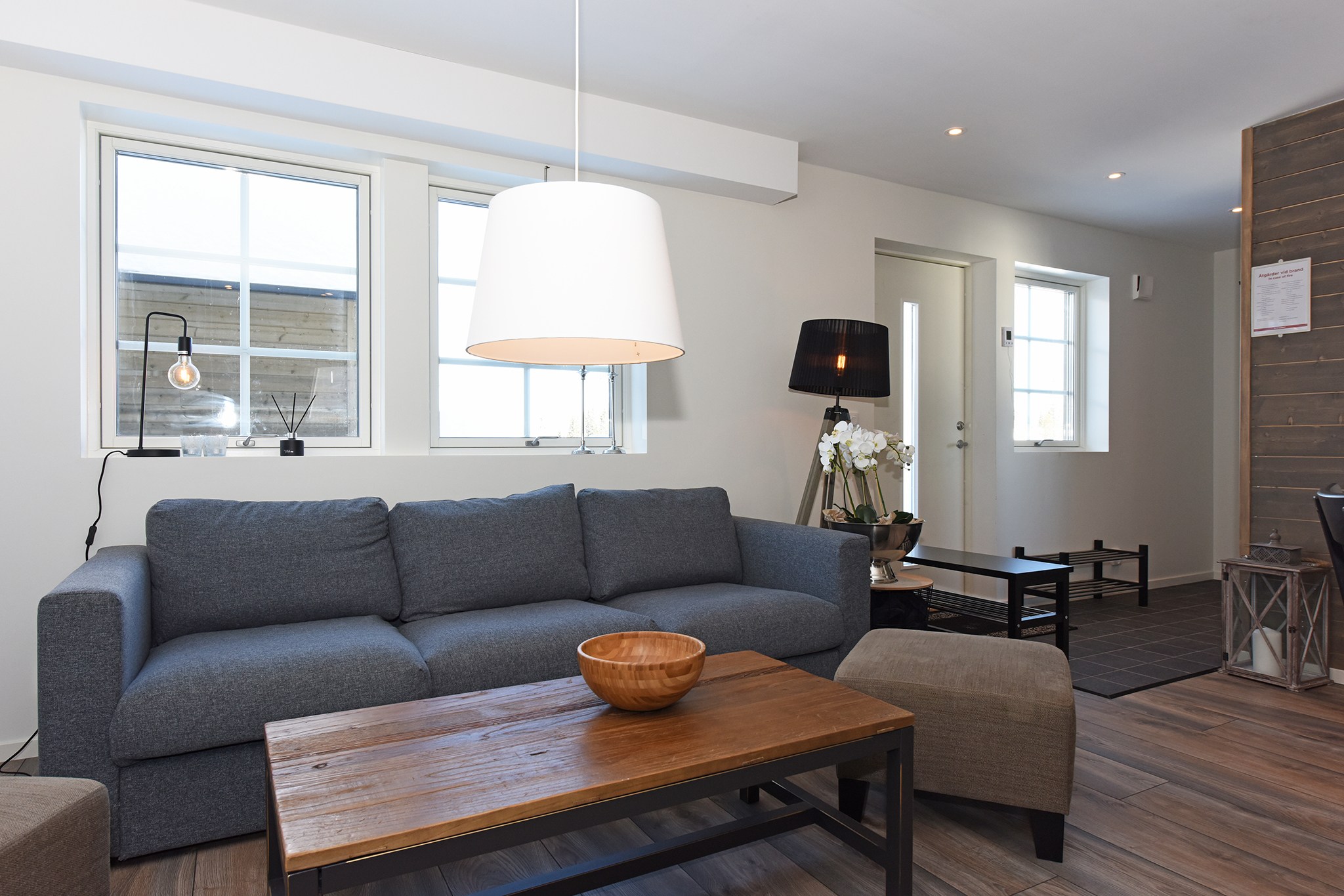 En moderne stue med en grå sofa, et sofabord i træ og en beige lænestol fremkalder den hyggelige atmosfære i Wagrain. En stor pendel hænger ovenover, med to vinduer, der tillader naturligt lys at strømme ind som en østrigsk sommerferie. I baggrunden fuldender en gulvlampe og dekorative genstande scenen.