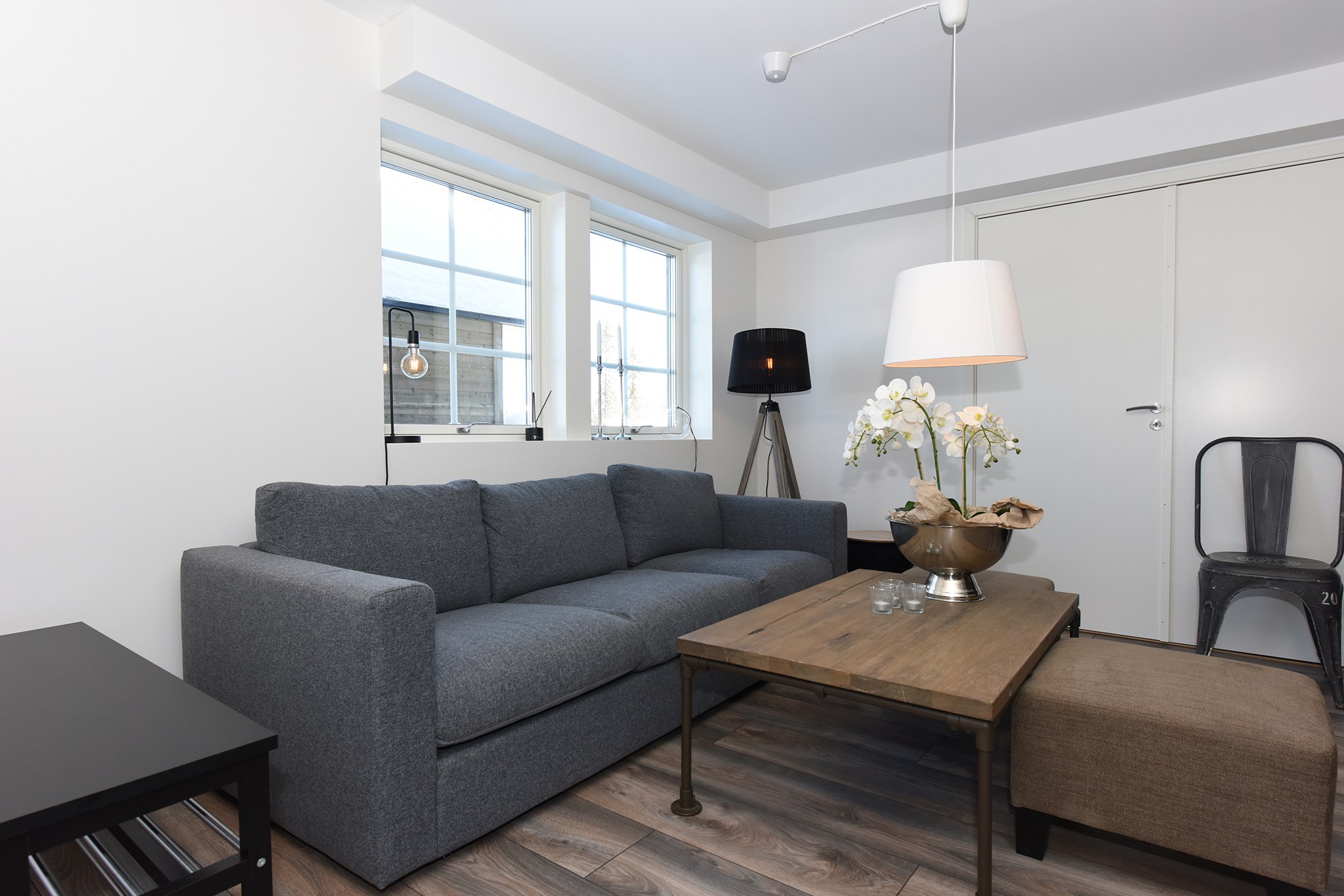 En moderne stue med en grå sofa, et sofabord i træ og en sort gulvlampe fanger elegant essensen af Westendorfs elegance. En hvid orkidé i en vase pryder bordet. Store vinduer, en beige skammel og en metalstol skaber en minimalistisk og hyggelig atmosfære, der minder om sommerferie Østrig.