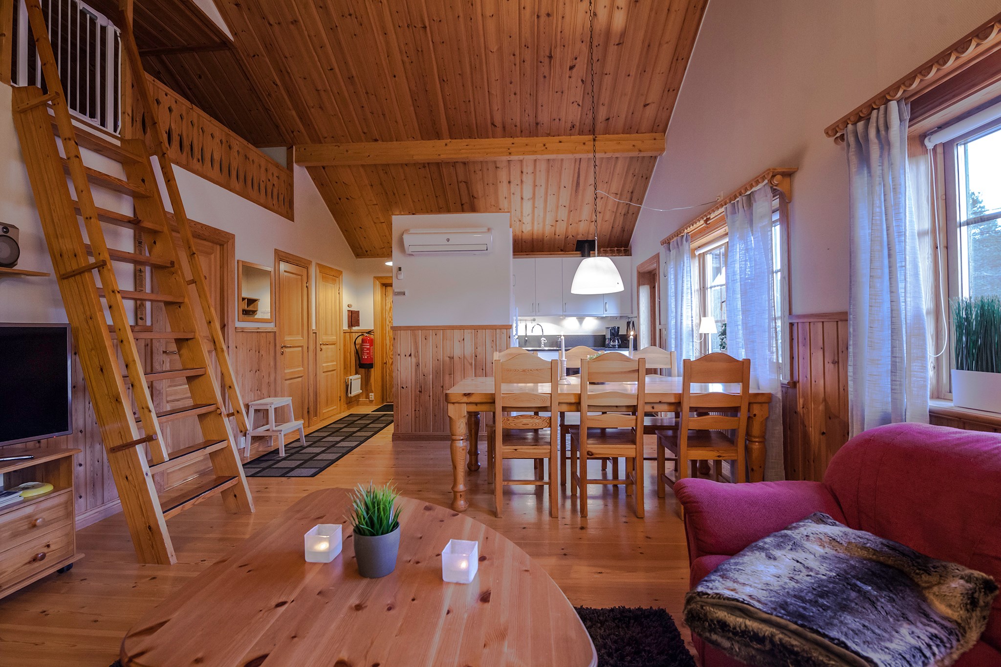 Et hyggeligt træinteriør har et rustikt spise- og opholdsområde i åbent plan, perfekt til at slappe af efter en dag på skiferie i Saalbach. Det omfatter et spisebord i træ med stole, en rød lænestol og en stige, der fører til en hems. Naturligt lys strømmer gennem store vinduer, og stearinlys pynter på et lille bord.