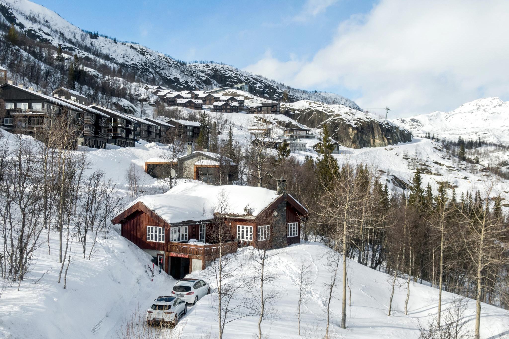 Et naturskønt vinterlandskab i Saalbach byder på en hyggelig træhytte omgivet af snedækkede træer og bakker. Flere andre kahytter er synlige i baggrunden, alle dækket af hvidt. To biler er parkeret foran hovedhytten, der antyder sommereventyr i sommerferie Østrig.
