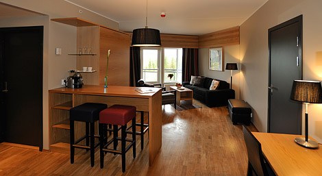En moderne stue med trægulve, en sort sofa, et lille sofabord og gulvlamper fremkalder charmen ved et Wagrain-retræte. En høj bordplade med to taburetter er i forgrunden. Store vinduespartier giver naturligt lys og giver en udsigt, der minder om sommerferie Østrigs frodige grønne områder.