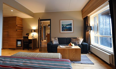Et moderne hotelværelse med et hyggeligt opholdsområde med en mørk sofa, et sofabord i træ og kunst på væggen. Perfekt til afslapning efter skirejser til Wagrain, lokalet er varmt oplyst, med et stort vindue til højre og en pæn opredt seng i forgrunden.
