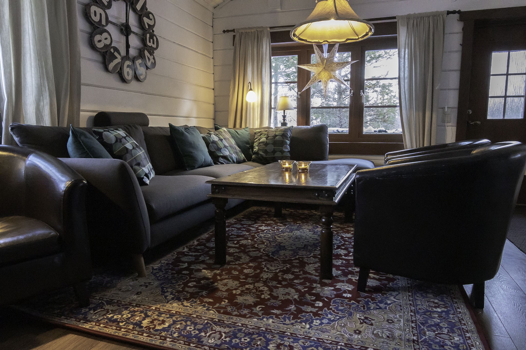 En hyggelig stue med en mørk sofa og lænestole omkring et sofabord i træ udstråler en Westendorf-charme. Et stort ur pryder væggen, og en stjerneformet lampe hænger ved et vindue med lys, der strømmer igennem. Gardiner indrammer udsigten, mens et mønstret tæppe dækker gulvet.