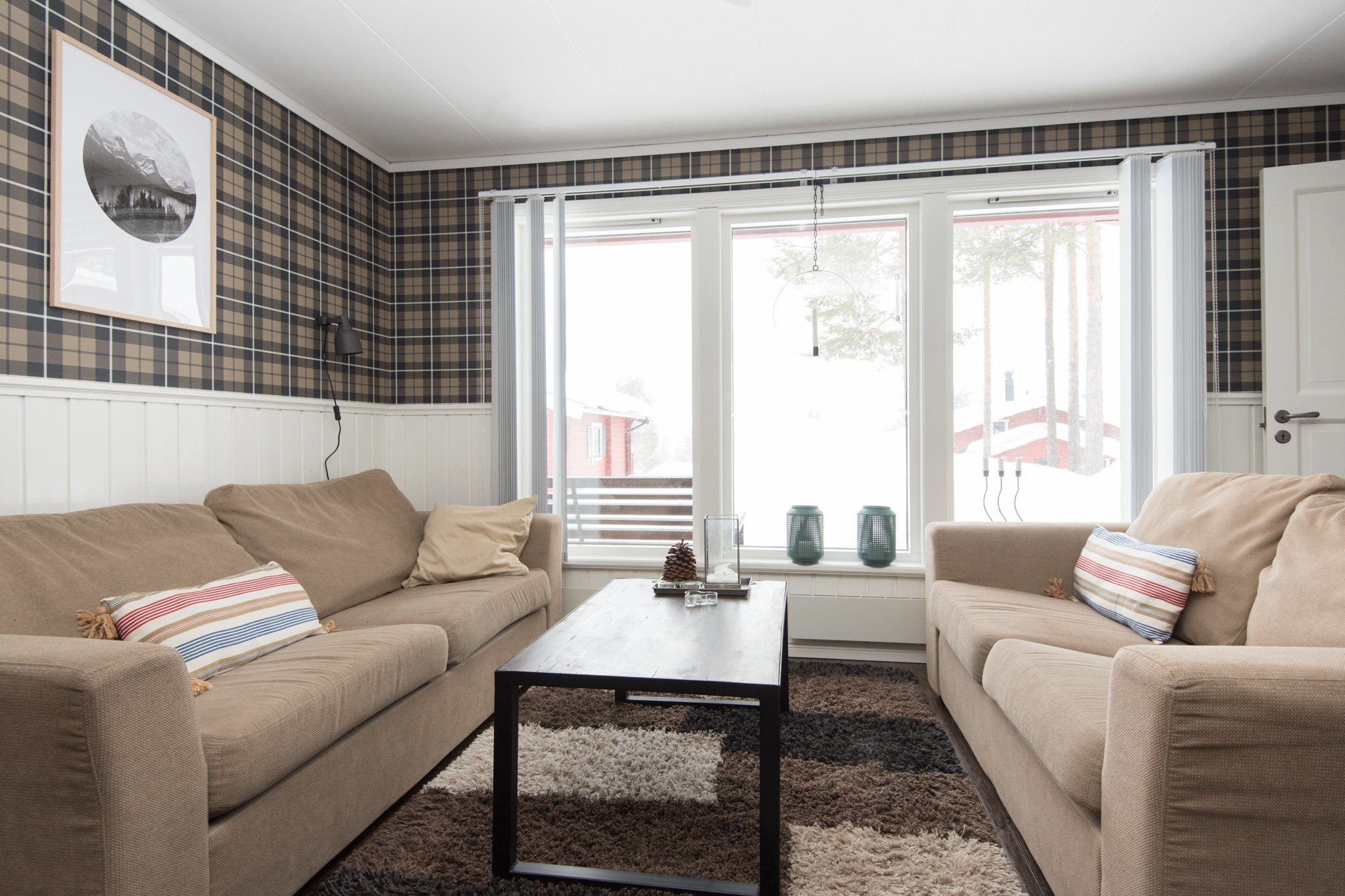 En hyggelig stue, der minder om en Saalbach skiferie, med to beige sofaer over for hinanden på tværs af et mørkt træ sofabord. Naturligt lys strømmer gennem rene gardiner på det store vindue, oplysende plaid tapet og et bjerglandskabsbillede hængende på den ene væg.