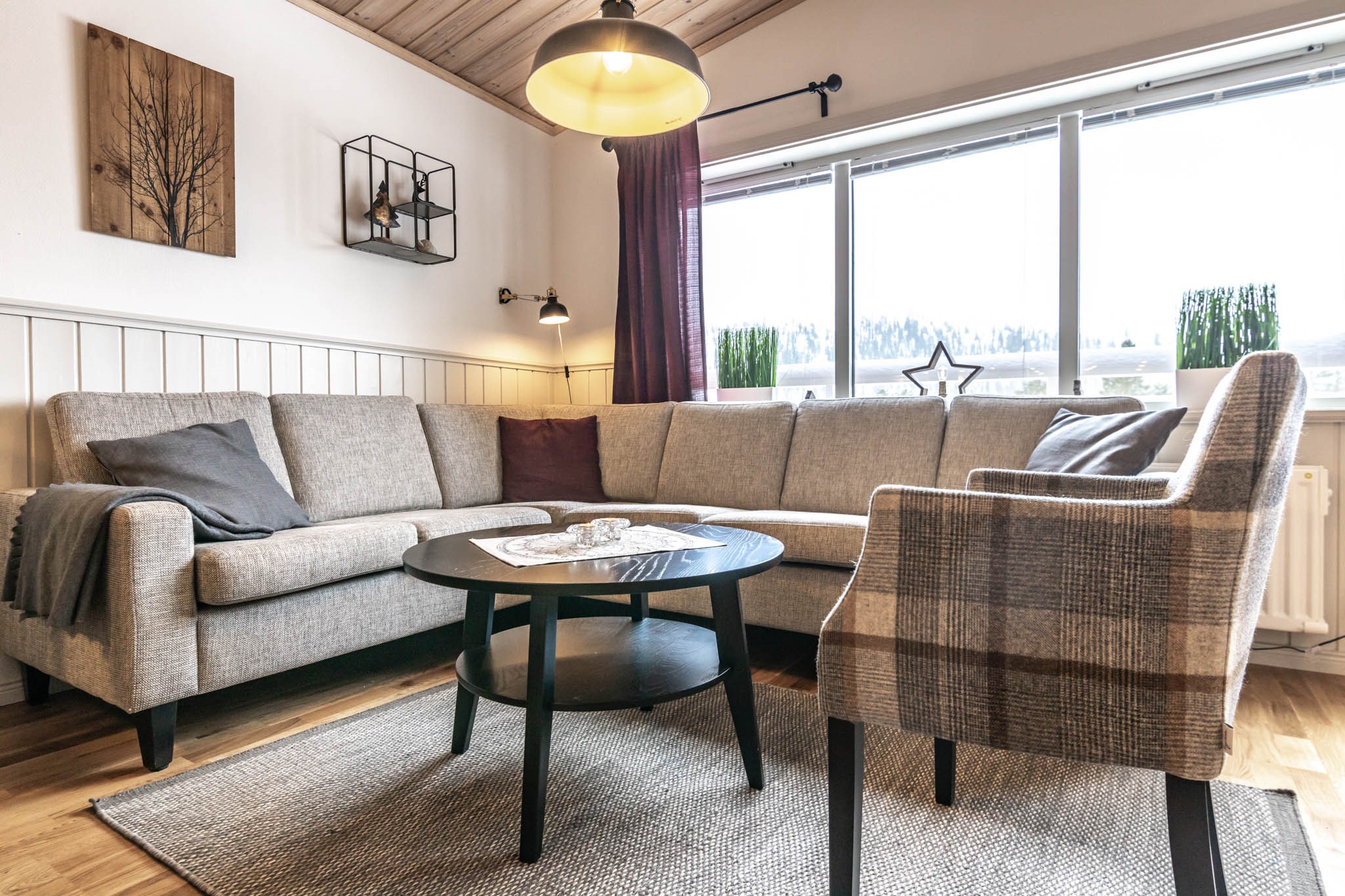 En hyggelig stue, der minder om et Westendorf-retræte, med en stor beige L-formet sofa, en plaid lænestol og et rundt sort sofabord på et gråt tæppe. Et vindue med lilla gardiner indrammer små planter på karmen. Vægindretningen inkluderer et alpint trækunstværk og en sort metalhylde.