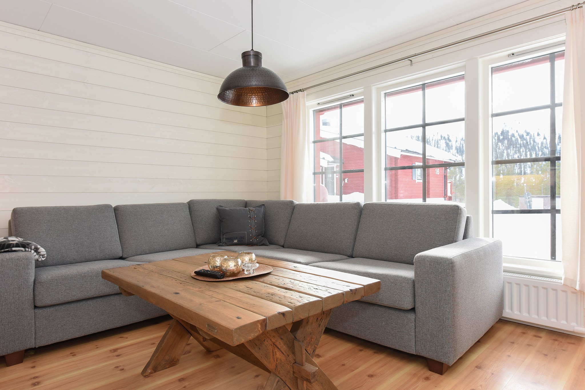 En hyggelig stue, der minder om en Westendorf-hytte, har en grå sektionssofa, et træsofabord prydet med dekorative genstande og et stort vindue, der bader rummet i naturligt lys. En pendel hænger elegant ovenover og komplementerer de lyse trægulve og hvide vægge.