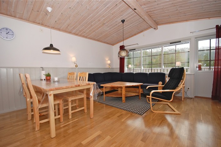 Et hyggeligt opholds- og spiseområde har et træbord med stole, en blå sektionssofa, en sort loungestol og to sofaborde. Et tæppe sidder i nærheden under moderne pendler, med store vinduer, der giver naturligt lys - et perfekt tilflugtssted, der minder om en Wagrain-skiferie.