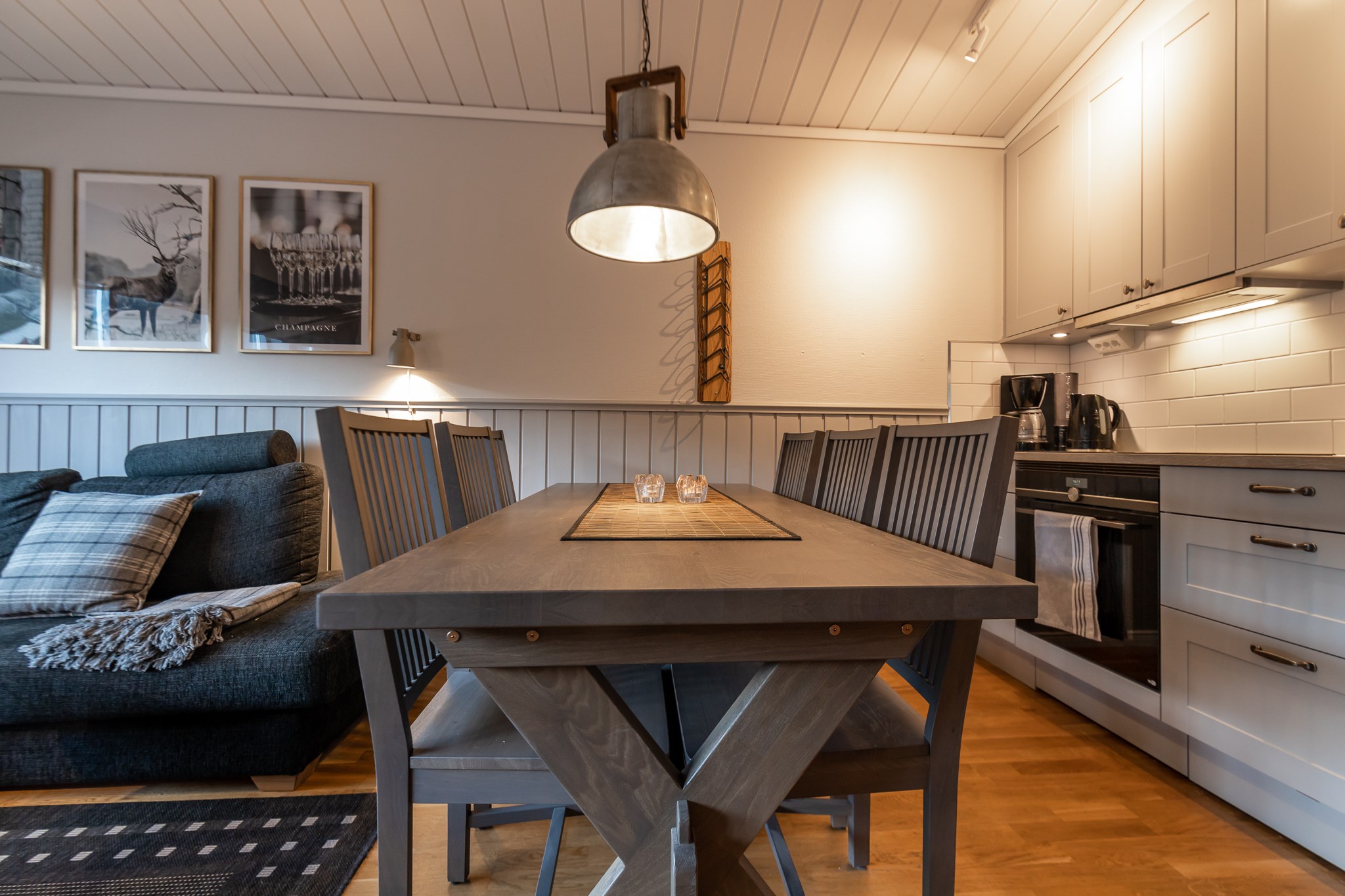 En hyggelig spiseplads med et træbord med en løber og seks stole. Til venstre står en mørkegrå sofa med plaidpuder, der minder om en skiferie i Westendorf, op ad en væg med indrammet kunst. Køkkenet er udstyret med lysegrå skabe og komfur, under varm, omgivende belysning.