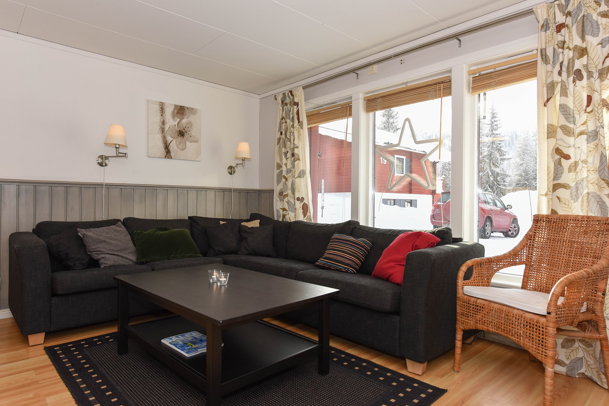 En hyggelig stue med en mørkegrå sektionssofa, et sort sofabord og en kurvestol. Store vinduer med blomstergardiner lukker naturligt lys ind og afslører en sneklædt scene udenfor, der minder om Saalbach, med en rød bil parkeret nær en rød bygning.