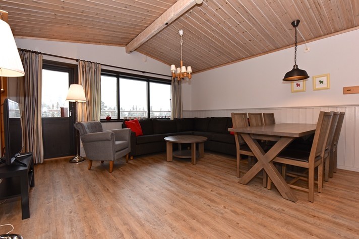 En hyggelig stue, der minder om en Westendorf-hytte, med træloft og gulv. Den har en lang sort sofa, en grå lænestol og et spisebord i træ med stole. To pendellamper hænger over bordet, mens et stort vindue bader rummet i naturligt lys.