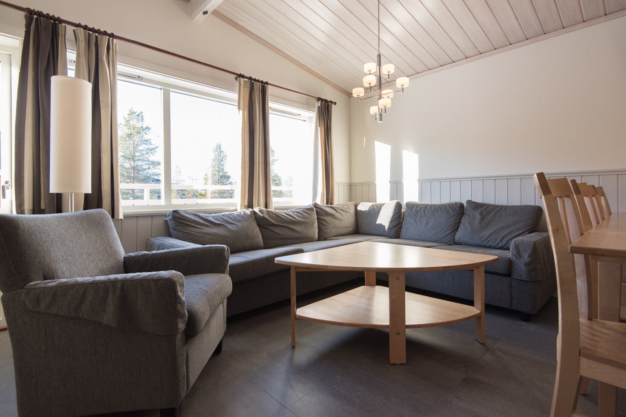 Hyggelig stue med stor grå sektionssofa og lænestol ud mod et sofabord i træ. Sollyset filtrerer gennem store vinduespartier med beige gardiner, der minder om en sommerferie Østrig atmosfære. Et spisebord af træ er synligt, og en moderne lysekrone hænger over hovedet.
