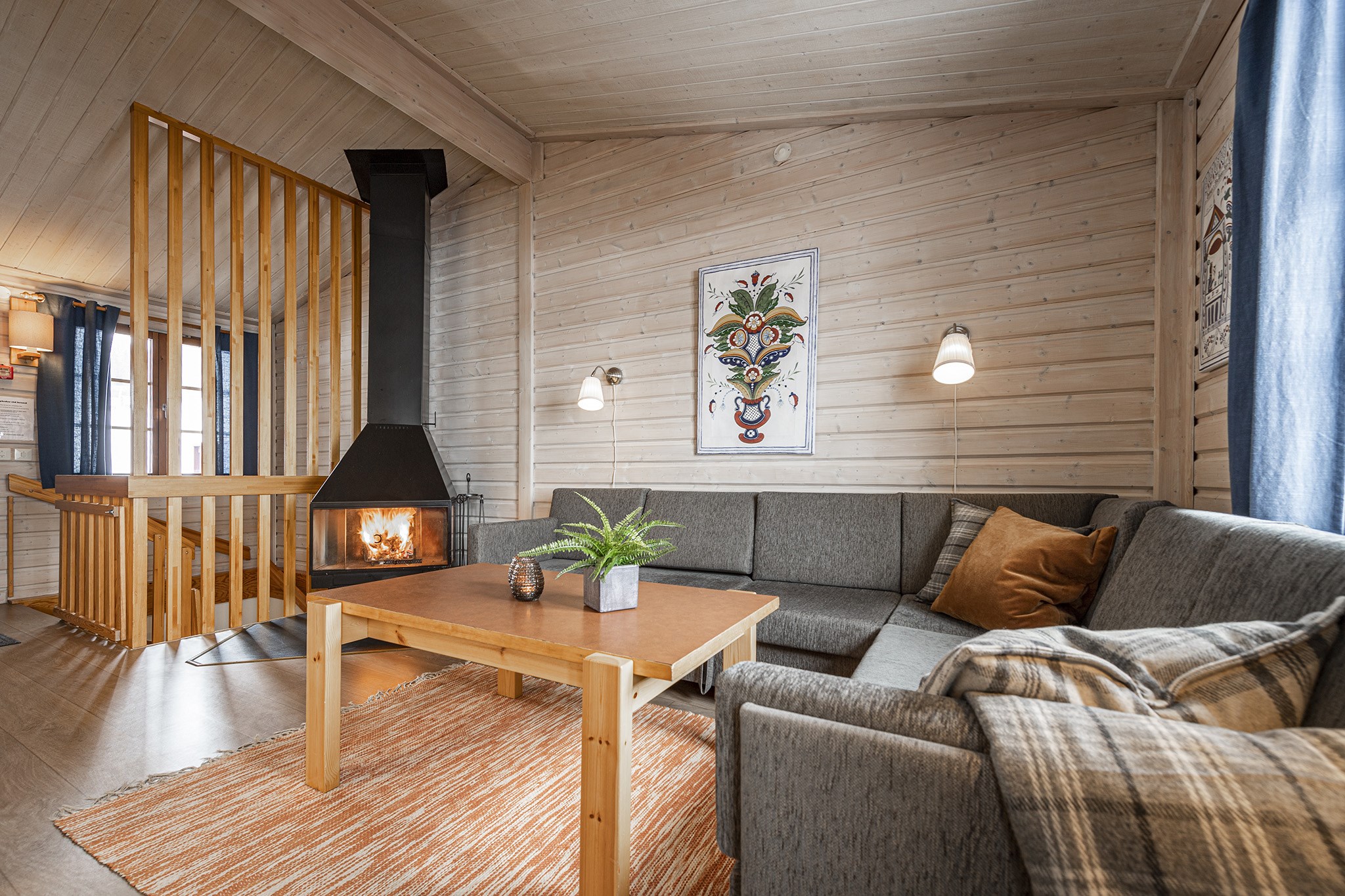 En hyggelig stue, der minder om en Saalbach skiferie, med trævægge og loft, har en grå L-formet sofa draperet i hynder og et tæppe. Et sofabord af træ hviler på et tekstureret tæppe, mens en moderne pejs varmer hjørnet under et dekorativt maleri.