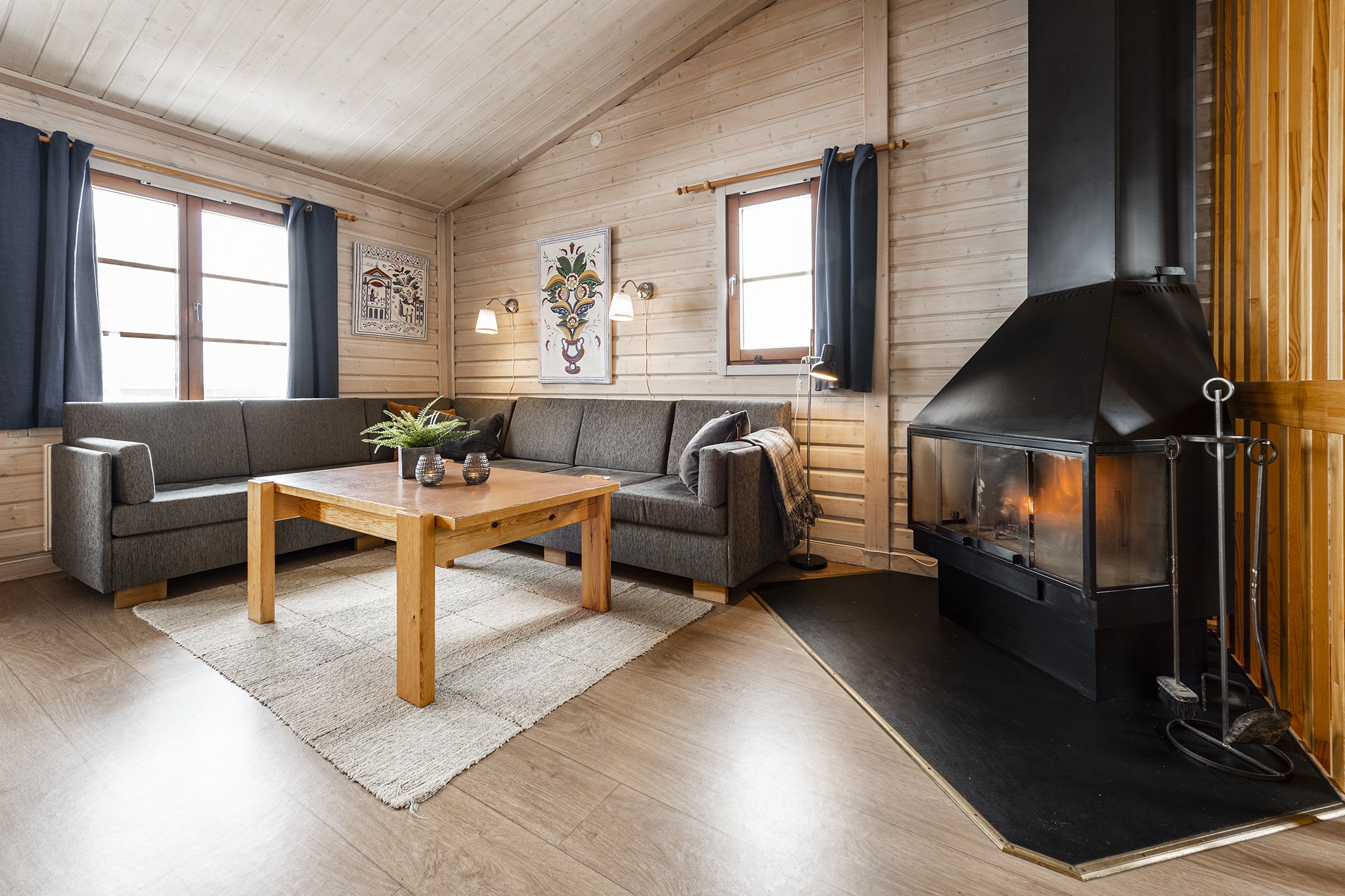 En hyggelig stue, der minder om et Westendorf-retræte, med trævægge og -loft, med en moderne sort pejs, en grå L-formet sofa med hynder og et sofabord i træ. Dekorativ kunst pryder væggene, når naturligt lys strømmer ind gennem to vinduer.