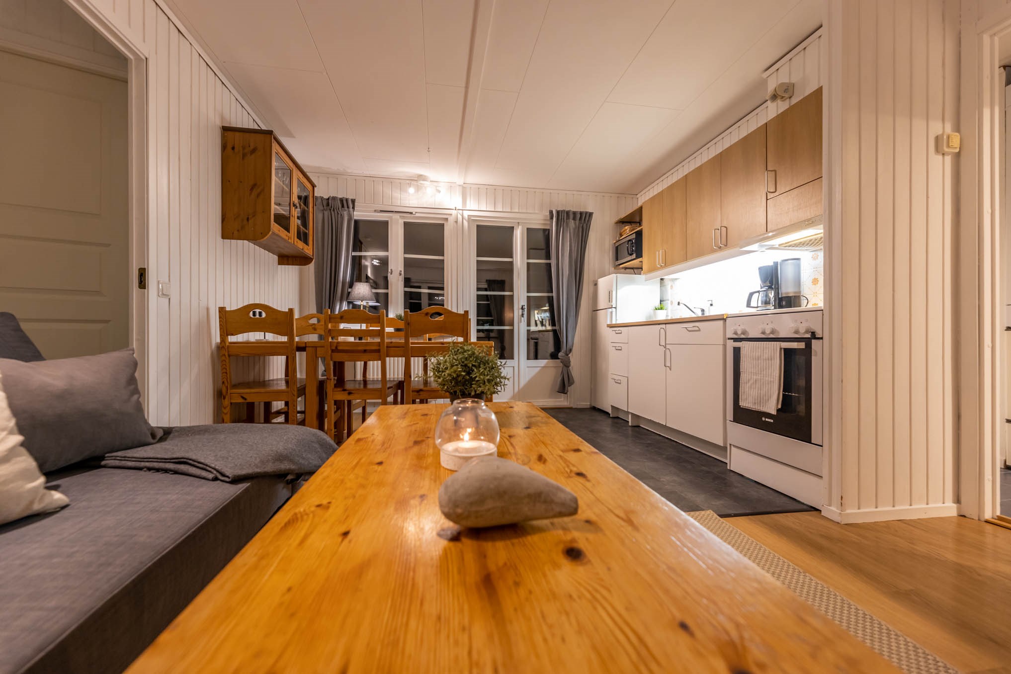 Hyggelig stue og køkken med træaccenter, perfekt til en skiferieferie. Har et spisebord i træ med stole, en grå sofa og et sofabord i træ, der er prydet med en lille sten og et stearinlys. Hvide skabe liner køkkenet, mens store vinduer med grå gardiner oplyser rummet.