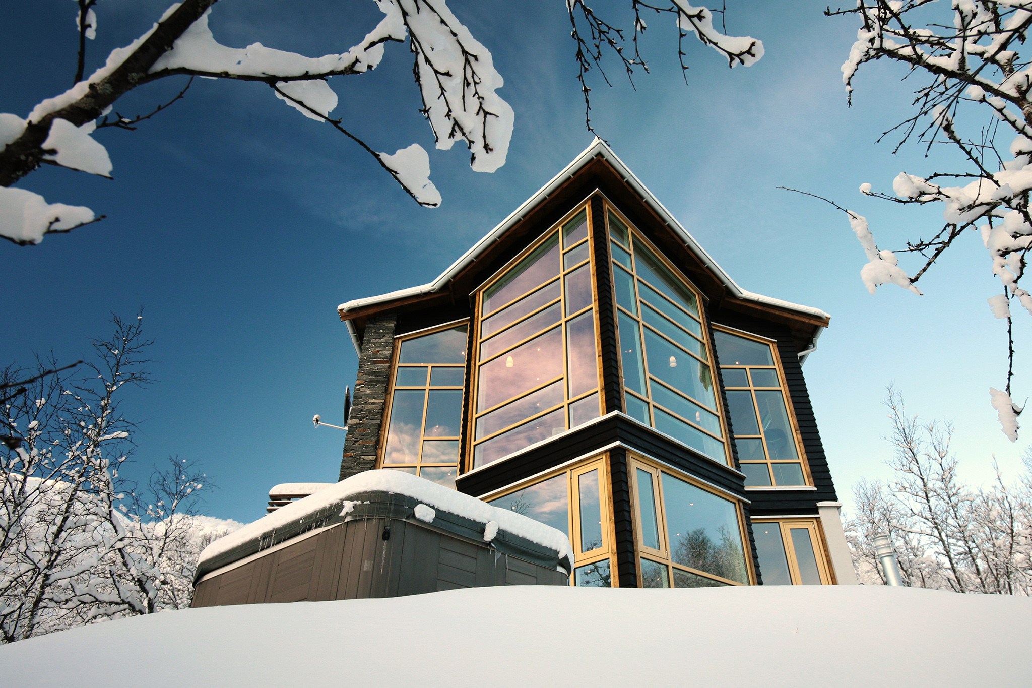En moderne hytte med glasfront omgivet af snedækkede træer i det fantastiske Saalbach. Bygningen har store vinduer og et stejlt, vinklet tag, sat mod en klar blå himmel og et snedækket landskab - ideel til uforglemmelige skirejser.