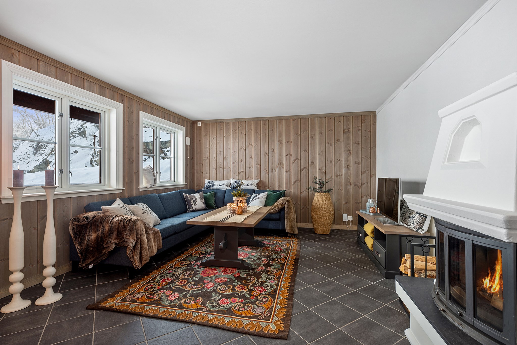 En hyggelig stue med en blå hjørnesofa, trævægge og et farverigt mønstret tæppe. En pejs med varm ild er til højre, og vinduer afslører et snedækket Westendorf-landskab. Perfekt til din skiferie flugt, rummet er dekoreret med stearinlys og puder.