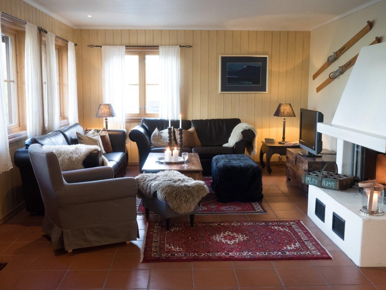 En hyggelig stue med sofaer og lænestole arrangeret omkring en hvid pejs, perfekt til at reflektere over tidligere skiferie. Et rødt mønstret tæppe dækker klinkegulvet, mens væghængte ski og et indrammet billede pryder de træbeklædte vægge. Blød belysning fra lamper skaber en varm stemning.