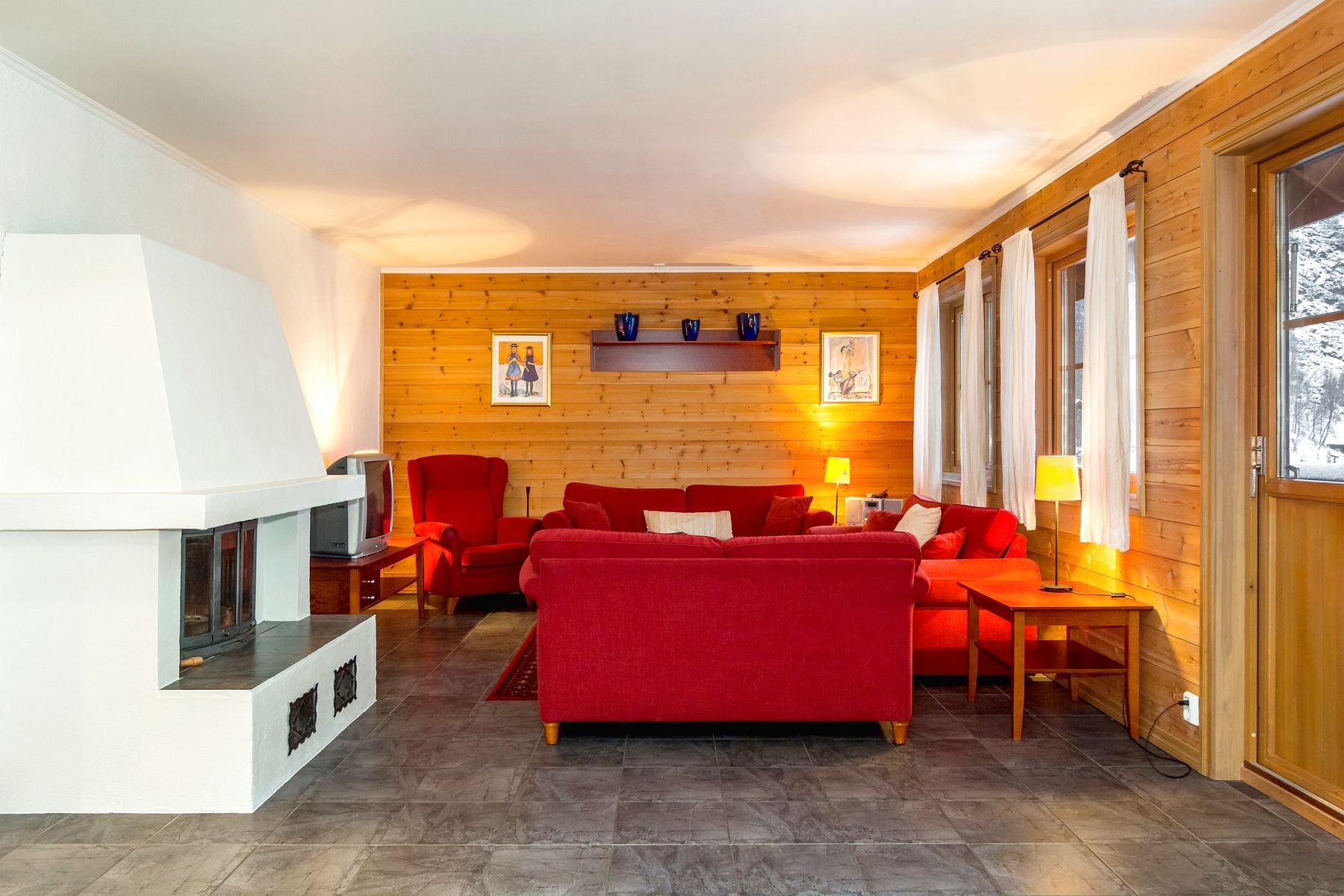Hyggelig stue i Westendorf med røde sofaer, trævægge og en hvid pejs. To lamper giver varm belysning, mens et tv sidder i hjørnet. Indrammet kunst og vaser pryder vægge og hylder. Et stort vindue og dør lukker naturligt lys ind, perfekt efter en dag med skirejser-eventyr.
