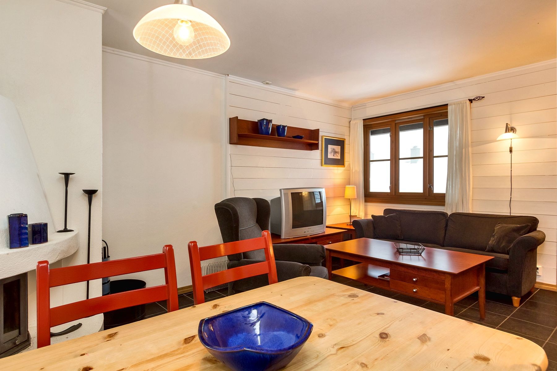 En hyggelig stue med hvide vægge har en sort sofa, et mørkt træsofabord, et vintage-tv og en pejs. Et spisebord i træ med røde stole er i forgrunden, der minder om charmerende Westendorf-interiør, mens en blå dekorativ skål er centreret på bordet.