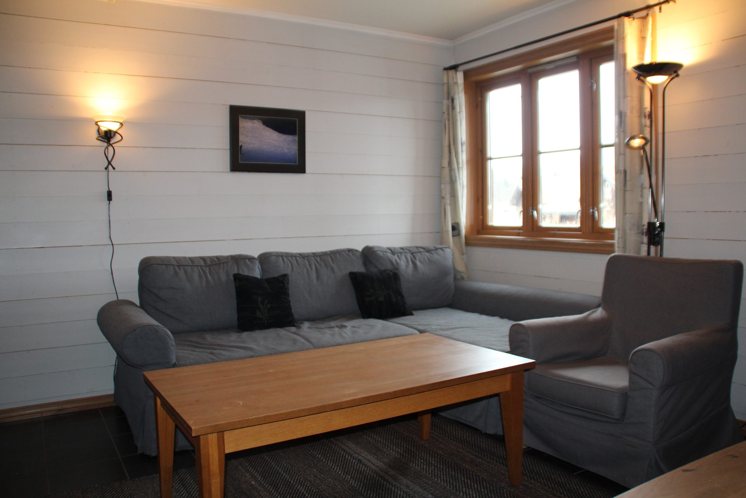En hyggelig stue i Westendorf, med en grå sektionssofa og matchende lænestol. Et sofabord i træ sidder foran. De hvide vægge rummer et indrammet billede over sofaen, mens vinduet med gardiner lukker naturligt lys ind, perfekt til planlægning af dine skiferieeventyr.