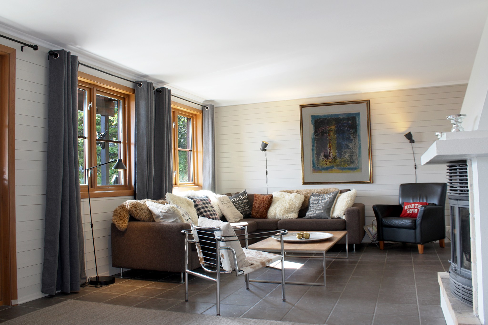 En hyggelig stue med en stor L-formet sofa prydet med hynder, en lækker stol og et lille sofabord. Trævinduer med grå gardiner indrammer udsigter, der minder om alpine retreater som Saalbach. Et moderne abstrakt maleri og en hvid pejs fuldender det indbydende rum.