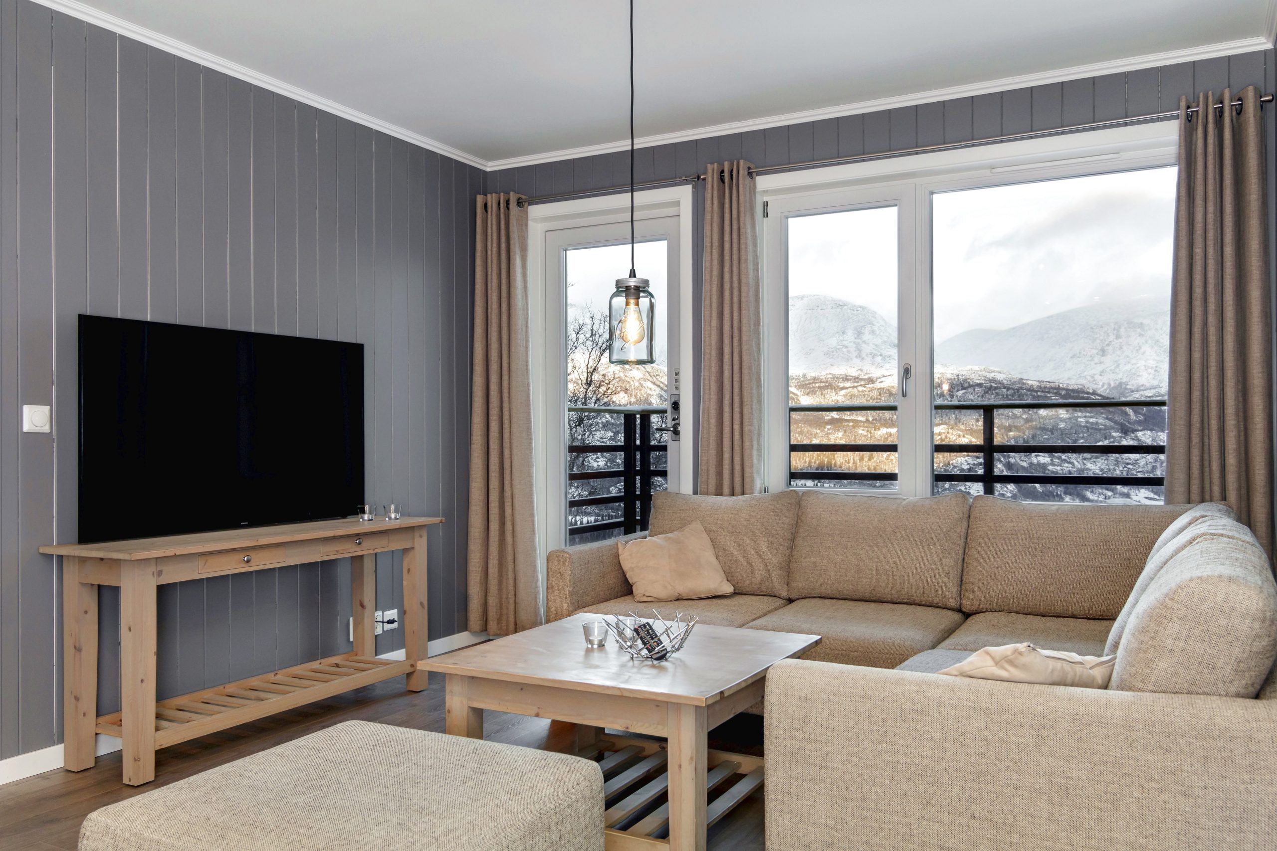 En hyggelig stue i Saalbach har en beige sofagruppe og et sofabord i træ. Et stort fladskærms-tv står på en træstativ, mens vinduerne, draperet med gardiner, afslører en sneklædt udsigt over bjergene. En pendel hænger elegant fra loftet og forstærker den alpine charme.
